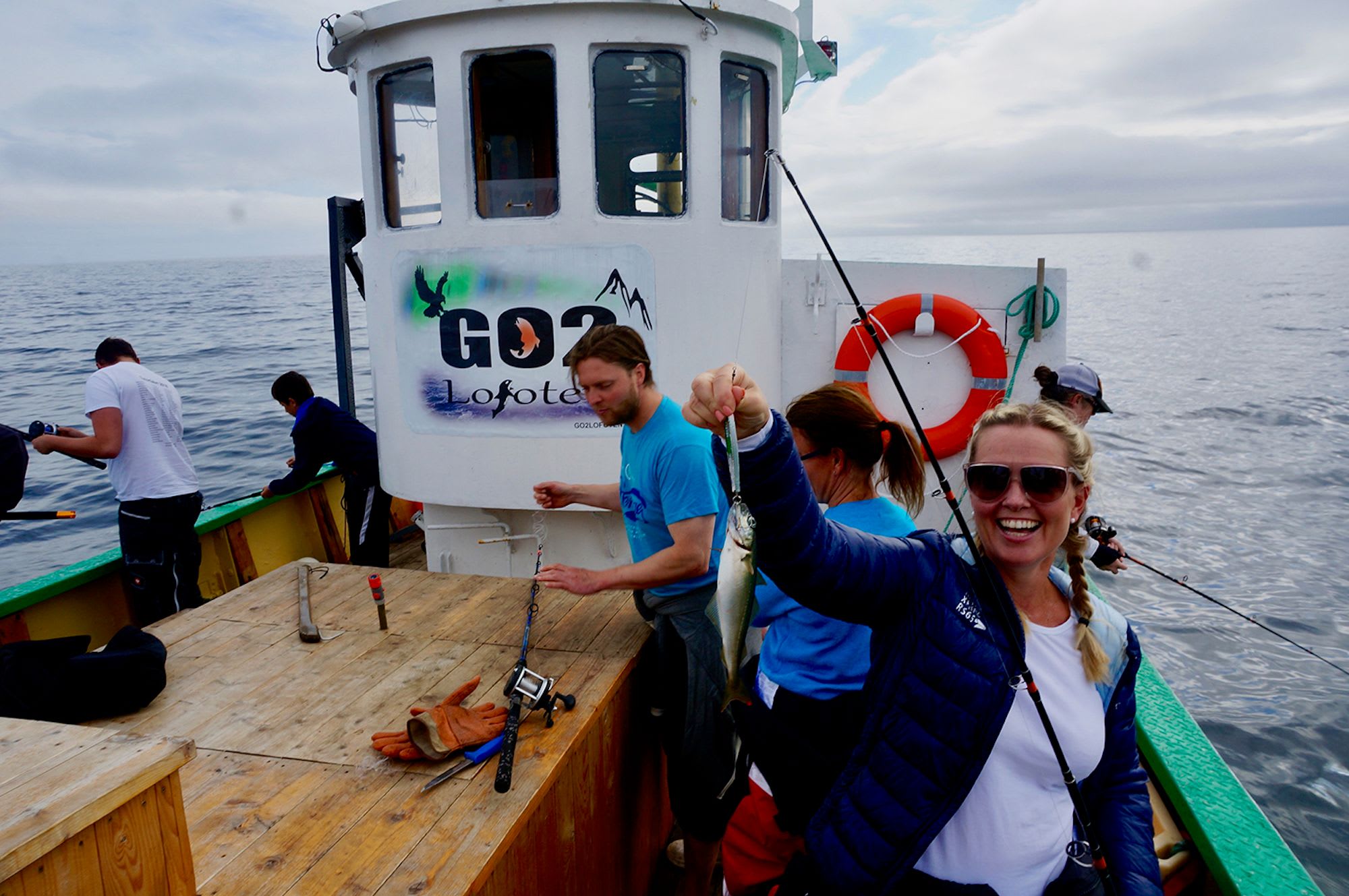 En kvinne holder opp fangsten sin, smilende, på en fiskebåt merket med "GO2 Lofoten"-logo.