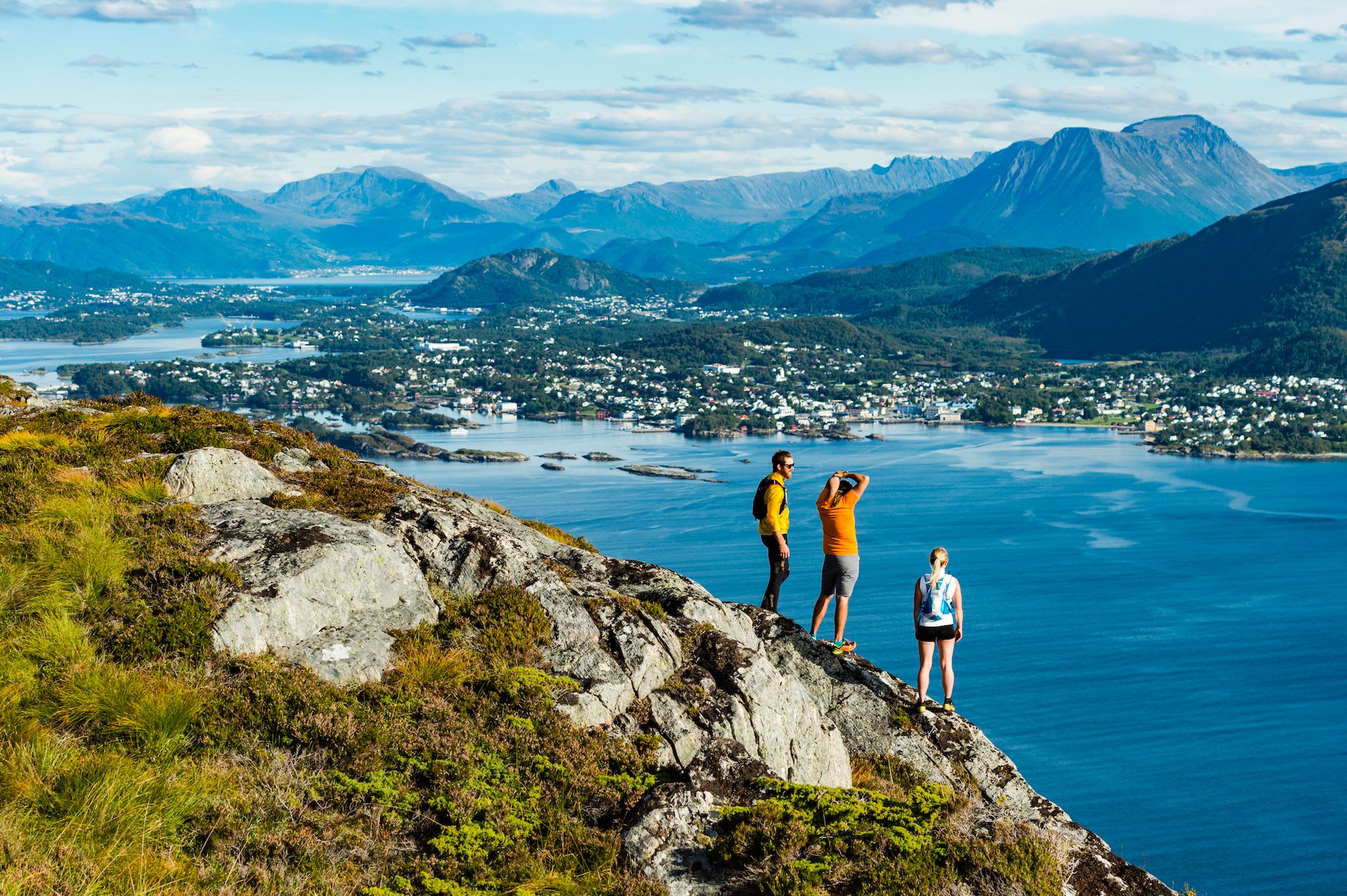 Tre turgåere står på toppen av et steinete fjell og nyter en panoramautsikt over byen, vannet og fjellene.