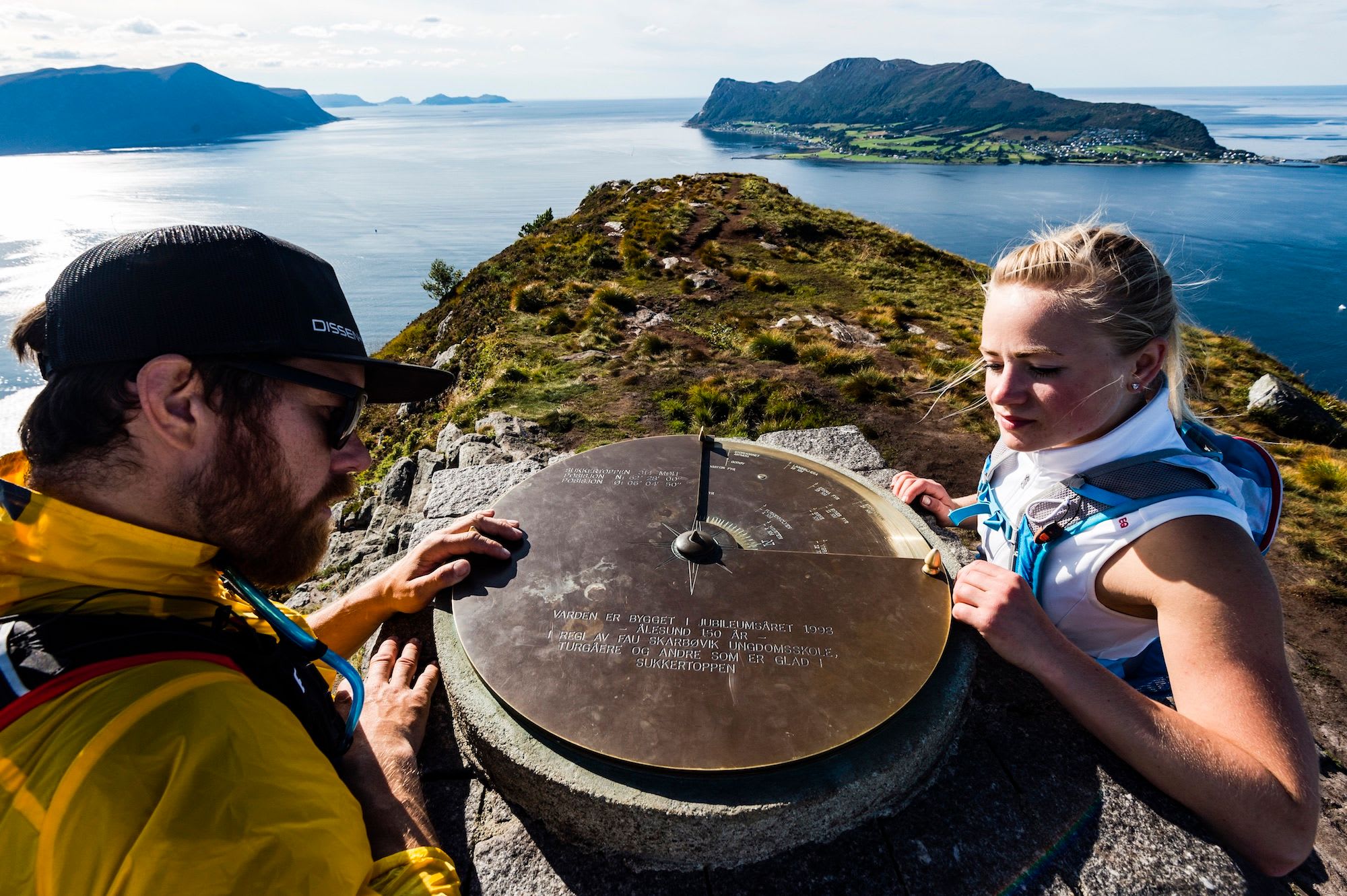 To turgåere undersøker et skilt på toppen av et fjell, med utsikt over kystlandskapet og byen nedenfor.