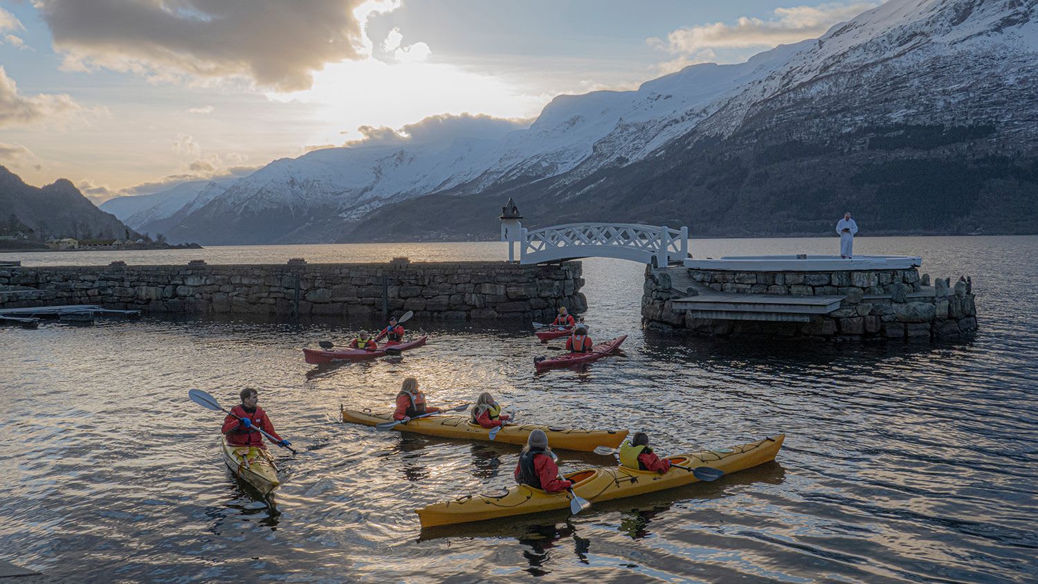 En gruppe kajakkpadlere padler på rolig vann, med en steinbrygge og snødekte fjell i bakgrunnen.