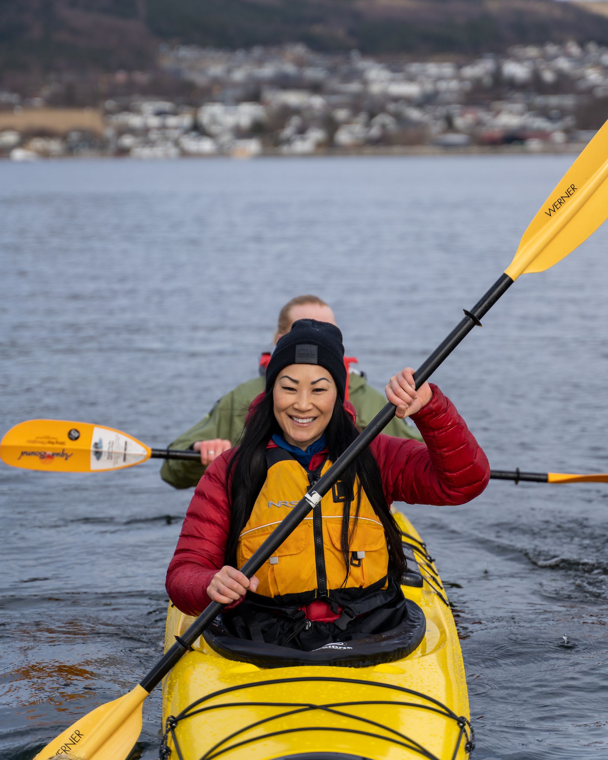 Nærbilde av smilende kvinne foran gul kajakk, by i bakgrunnen.