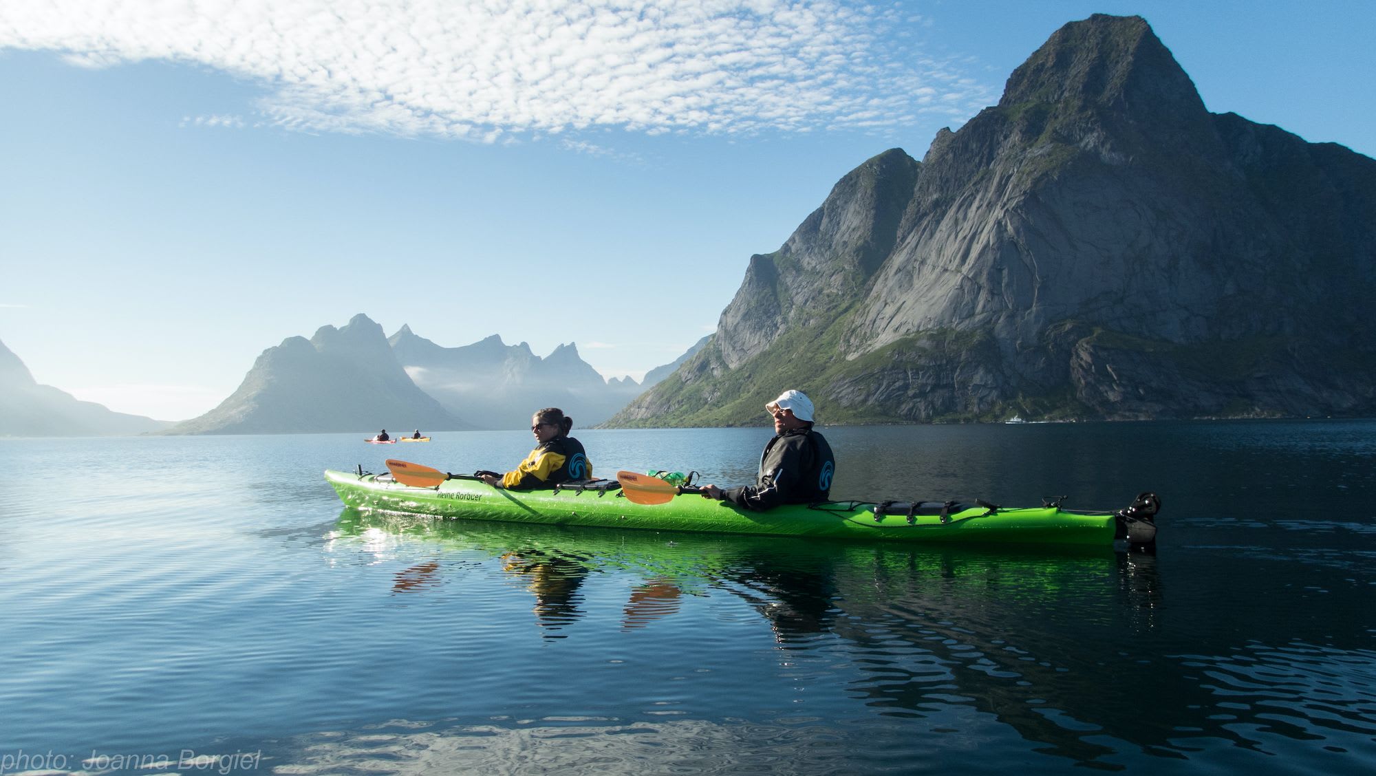 Two kayakers paddling on calm water with towering mountains in the background, under a clear sky with wispy clouds.