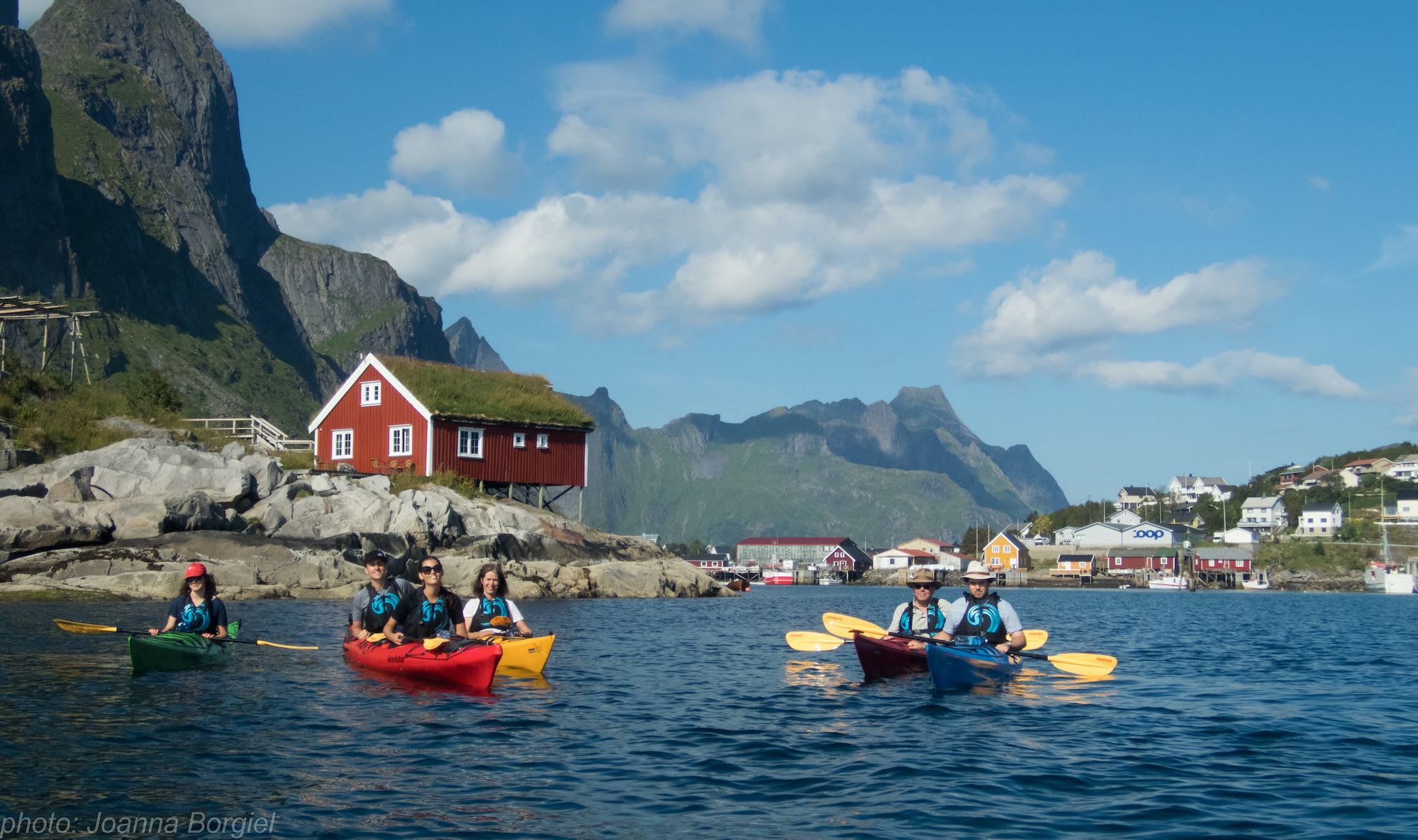 Kayakers paddling near a small island with a red house, green mountains, and blue sky, creating a peaceful and adventurous scene.