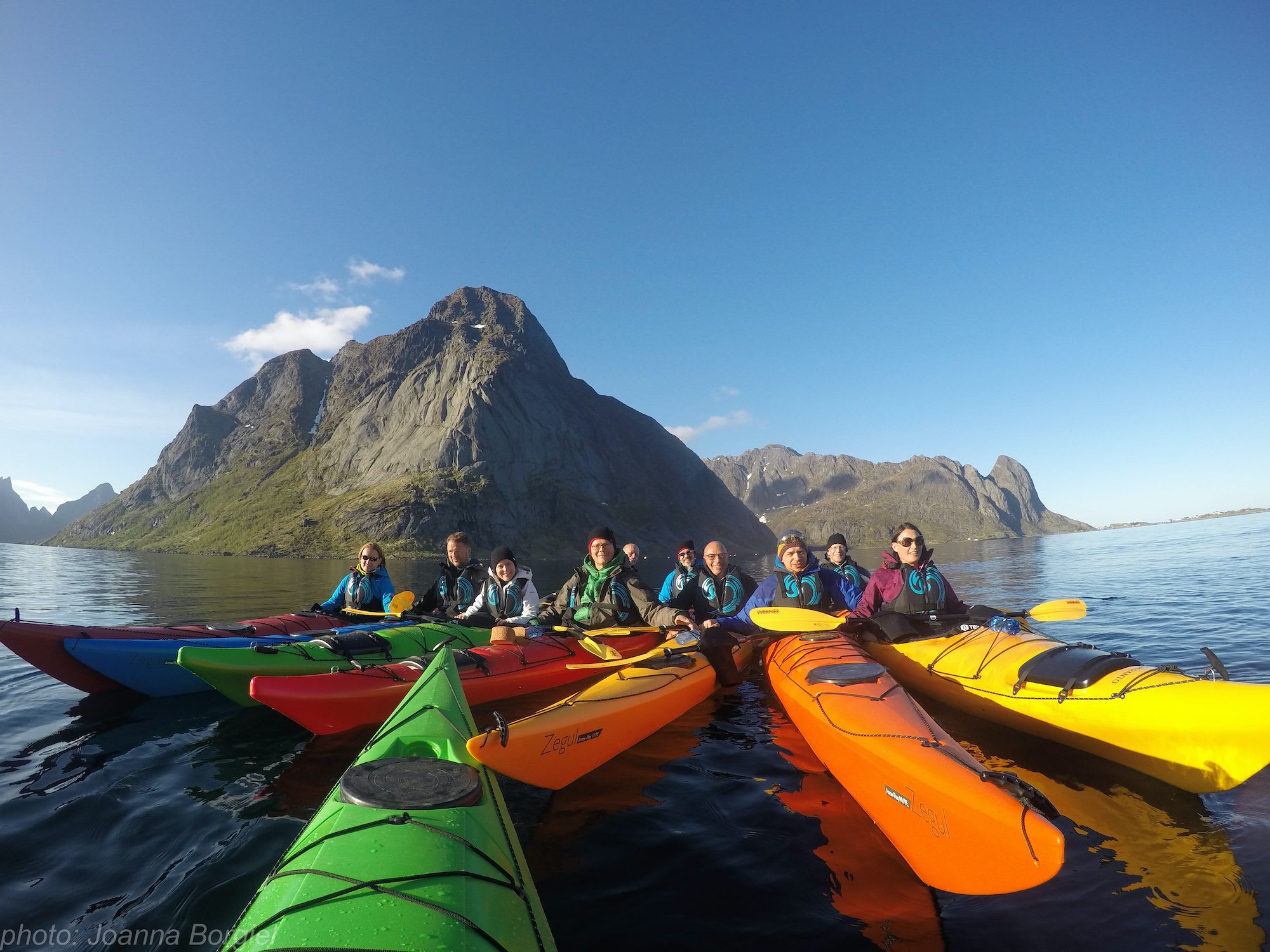 Kayakers gathered in colorful kayaks, smiling on calm, clear water with dramatic rocky mountains in the background.