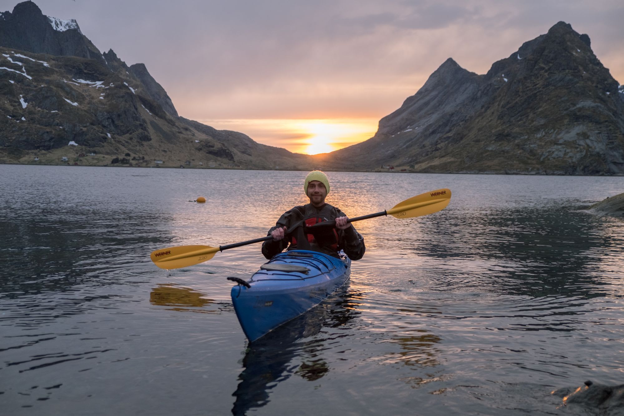 Person kayaking on a bright blue kayak with yellow paddles at sunset, with golden reflections on the water and rugged mountains in the background.