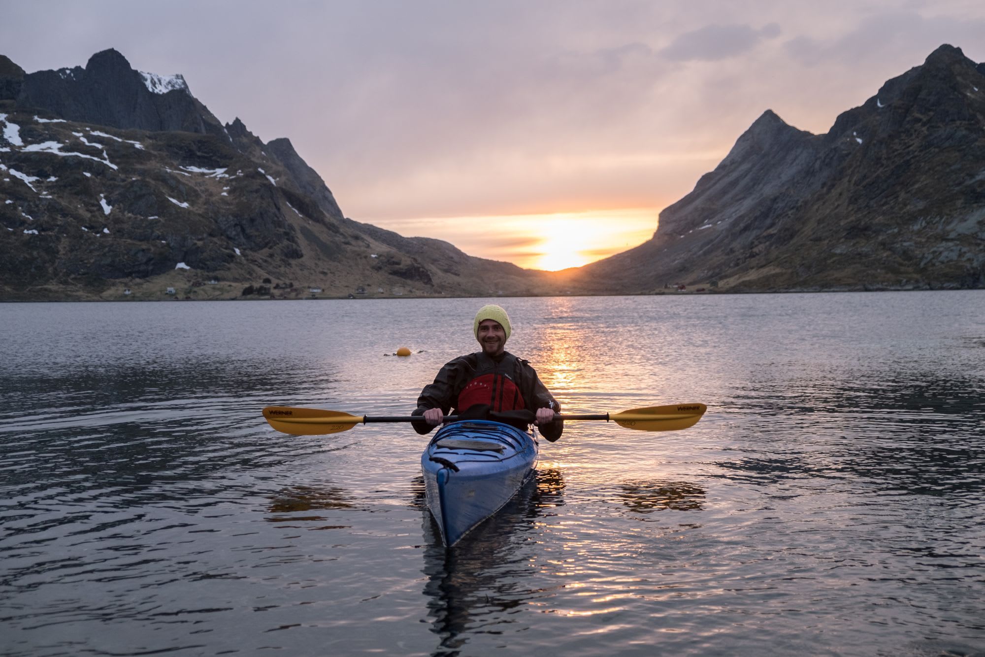 Smilende kajakkpadler i en blå kajakk som holder gule padler, på et rolig vann under solnedgang med gyldne refleksjoner på vannet.