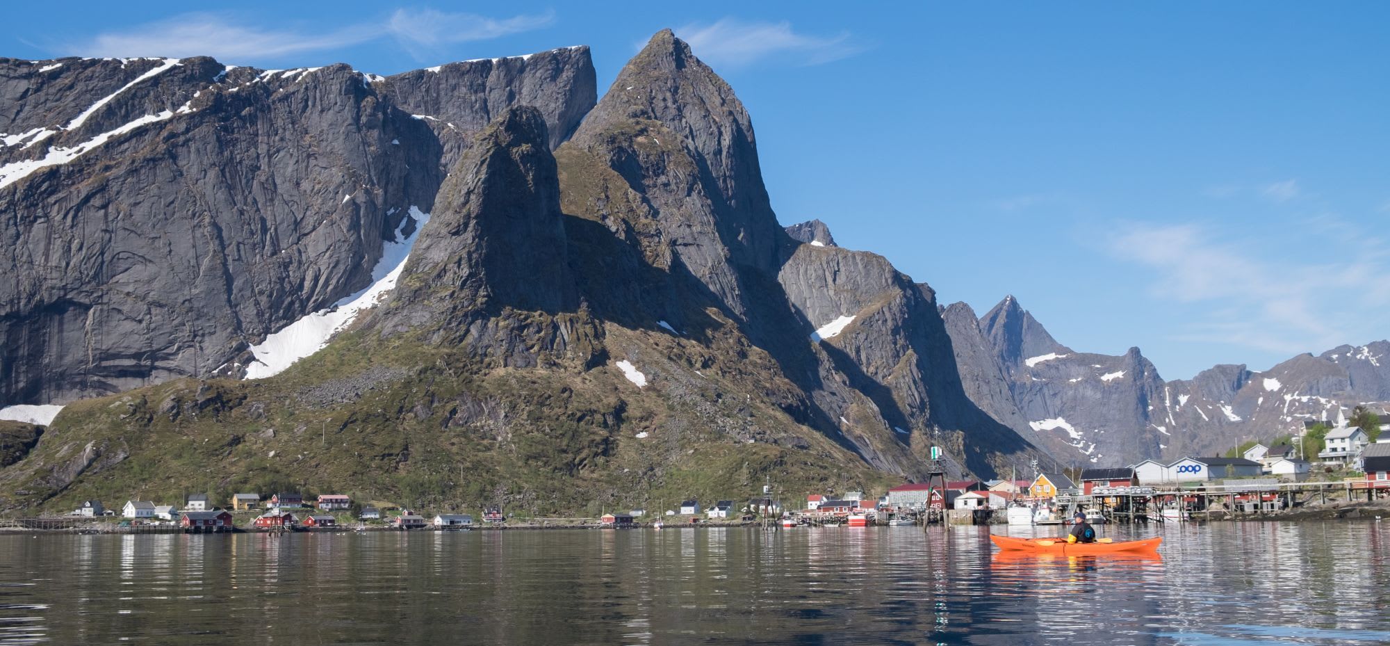 Person kayaking on a bright orange kayak on calm, reflective water near a coastal village with colorful houses and snowy mountains.
