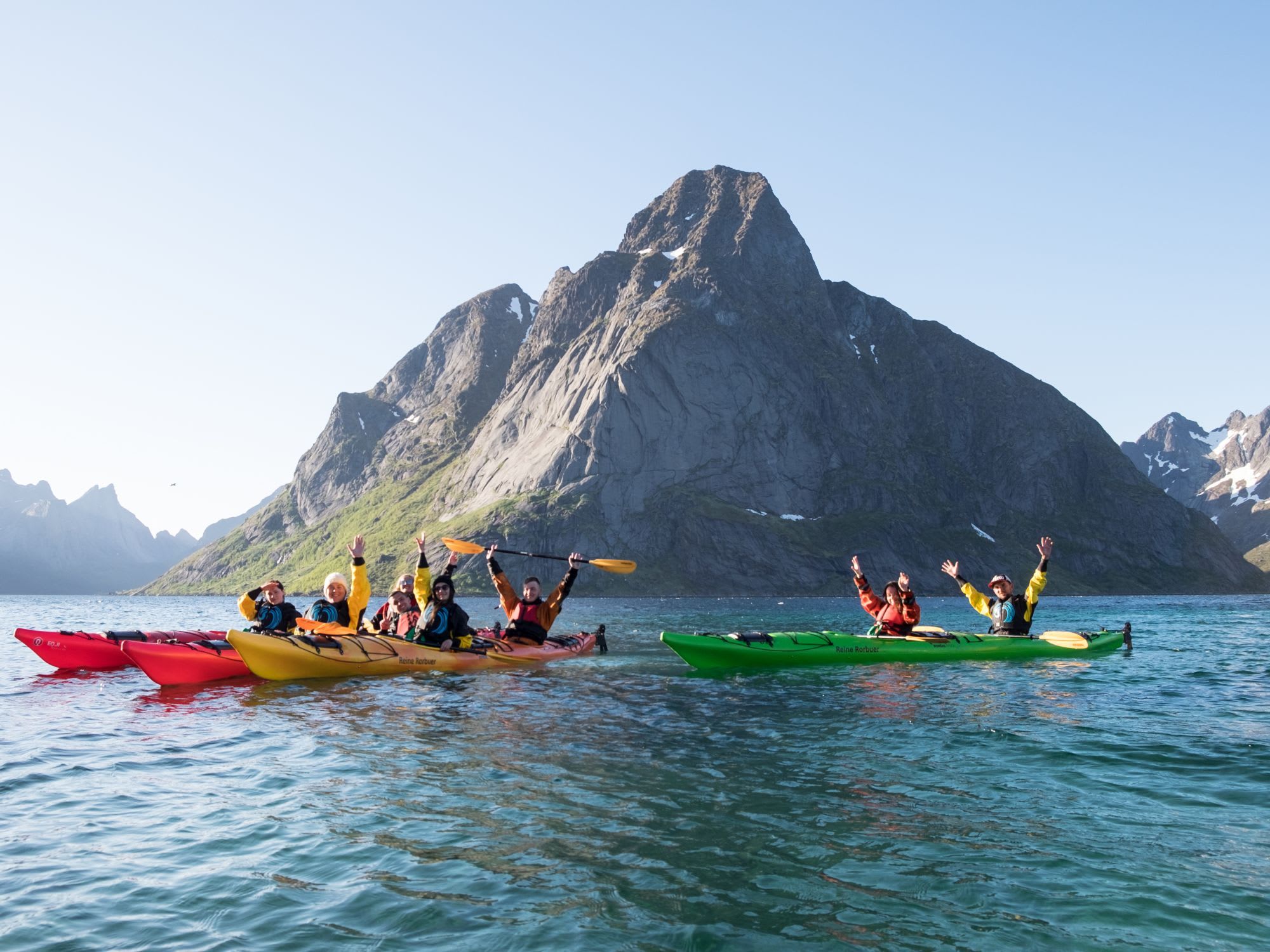 Group of kayakers in red, yellow, and green kayaks raising their arms in celebration on a turquoise lake with snowy mountains in the background.