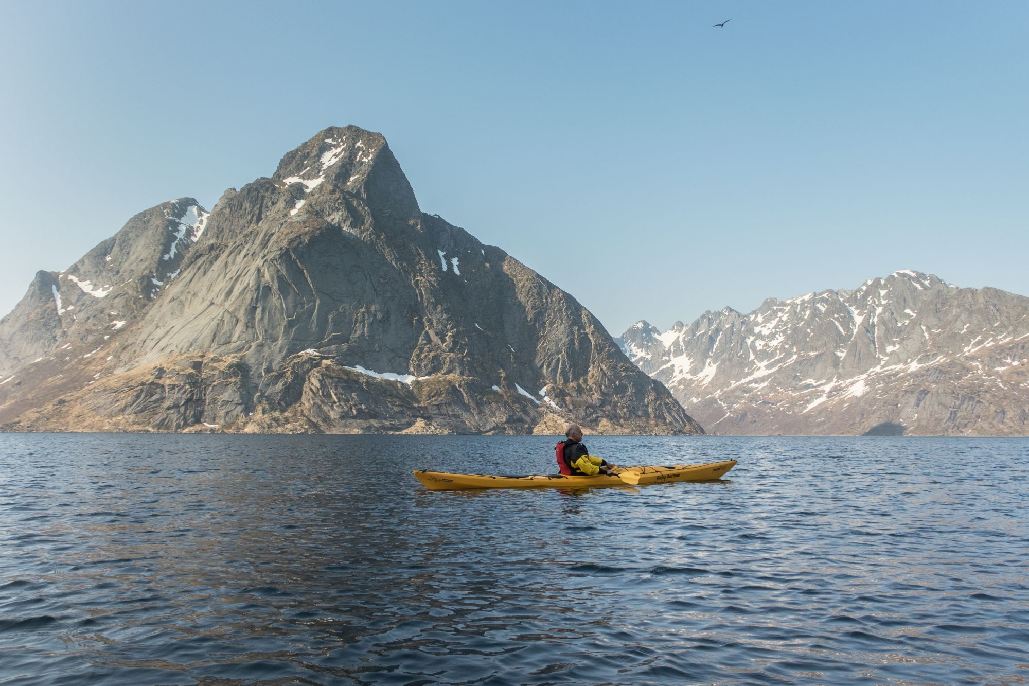 Person kayaking alone in a bright yellow kayak on a calm, dark blue lake, with towering snowy mountains and barren terrain in the background.