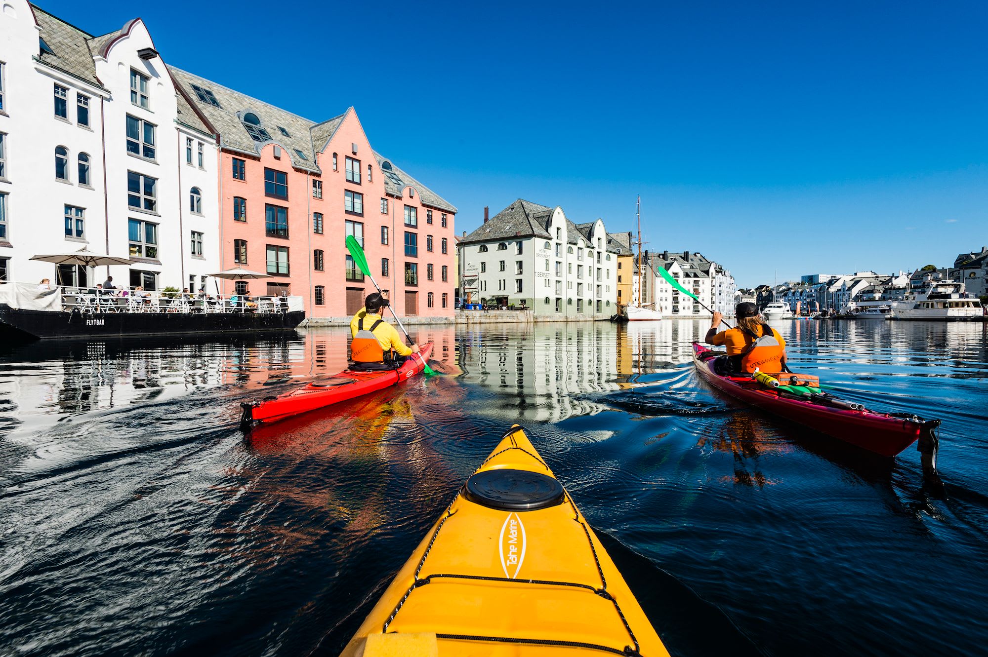 Two kayakers paddling red and yellow kayaks on calm water surrounded by colorful historic buildings, boats docked, and tourists enjoying the sunny day.