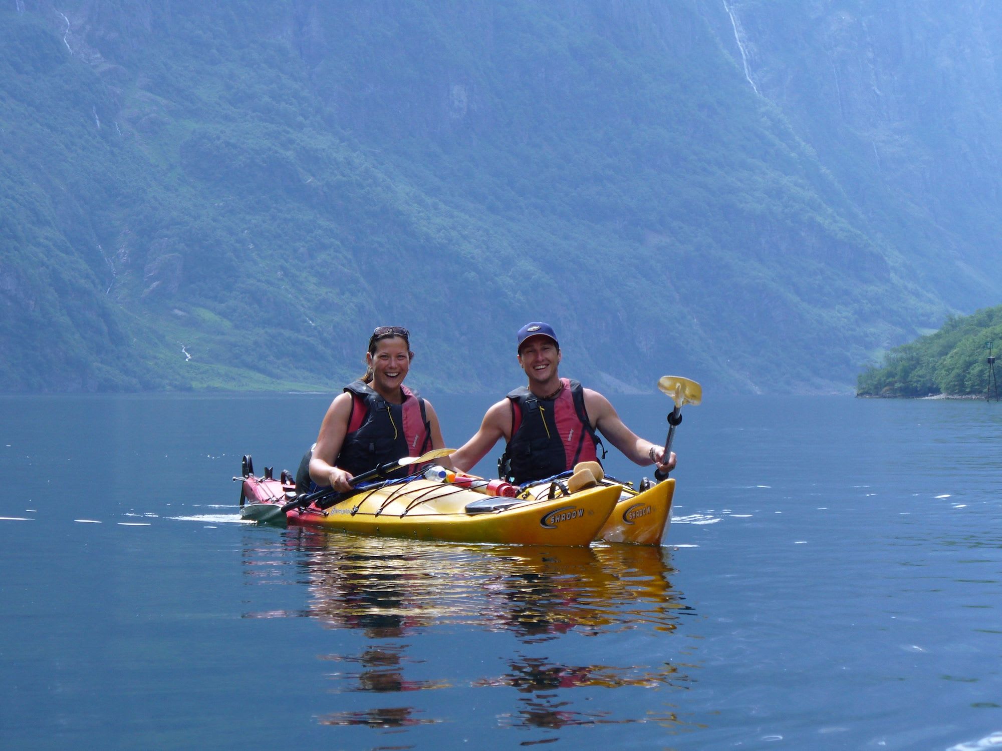 Two smiling kayakers in a yellow tandem kayak on calm blue water, surrounded by green hills and waterfalls in a peaceful fjord.