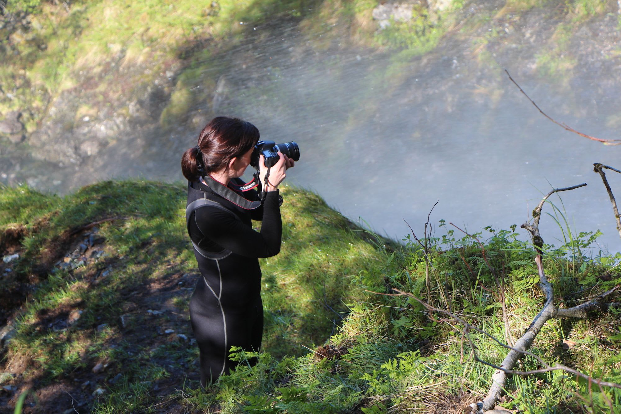 Woman in a black wetsuit standing on a moss-covered edge, taking photos with mist rising from the water below, surrounded by greenery.