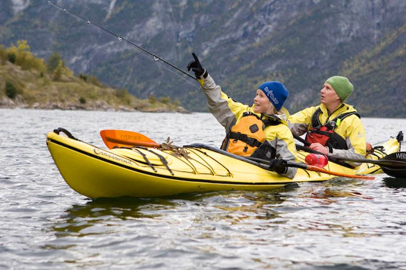To kajakkpadlere i en gul tandemkajakk padler på en rolig fjord, med en som peker entusiastisk, omringet av dramatiske fjell og høstfarger.