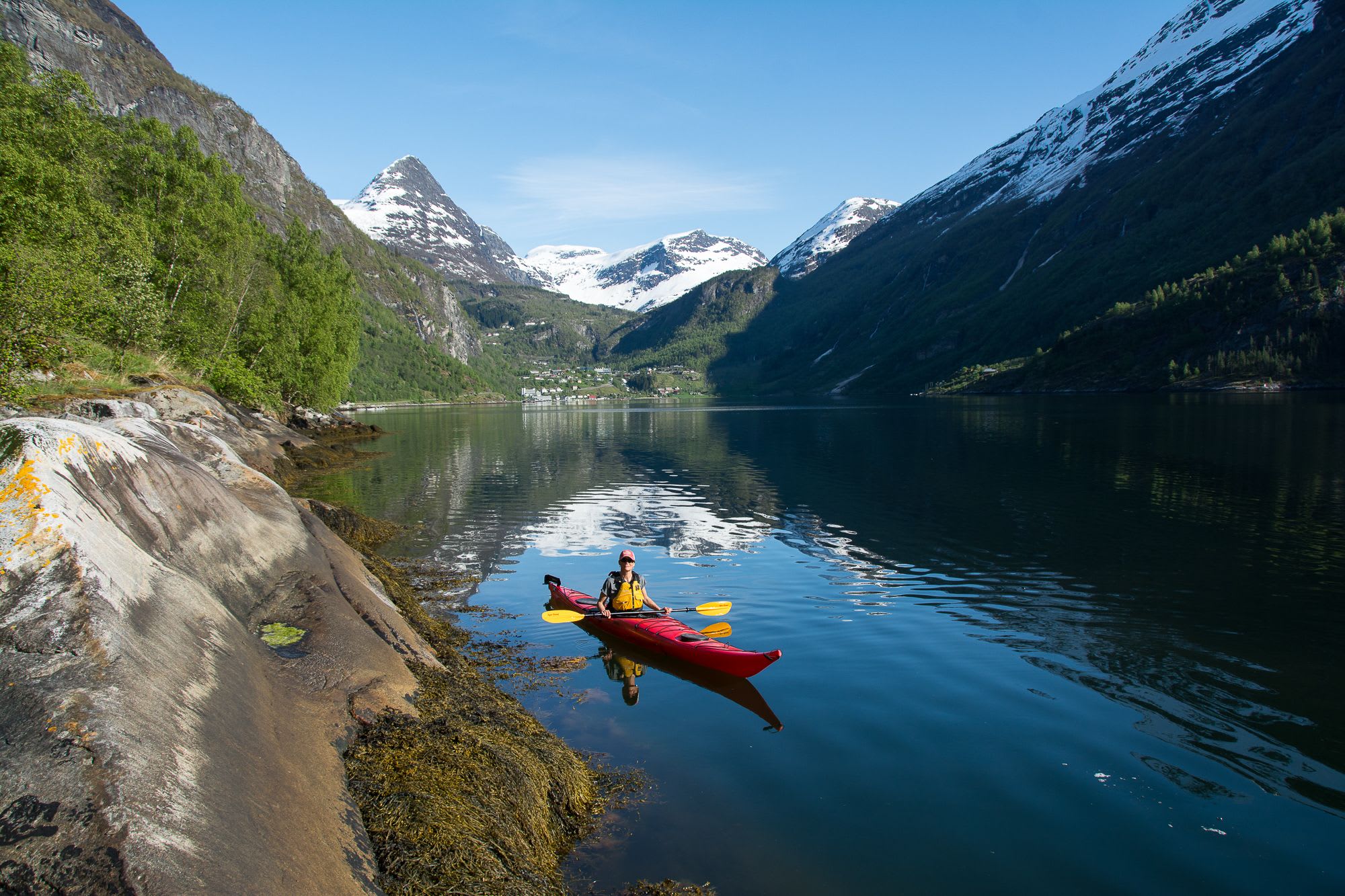 En person som padler i en rød kajakk på rolig vann, med snødekte fjell og en liten landsby synlig i bakgrunnen.