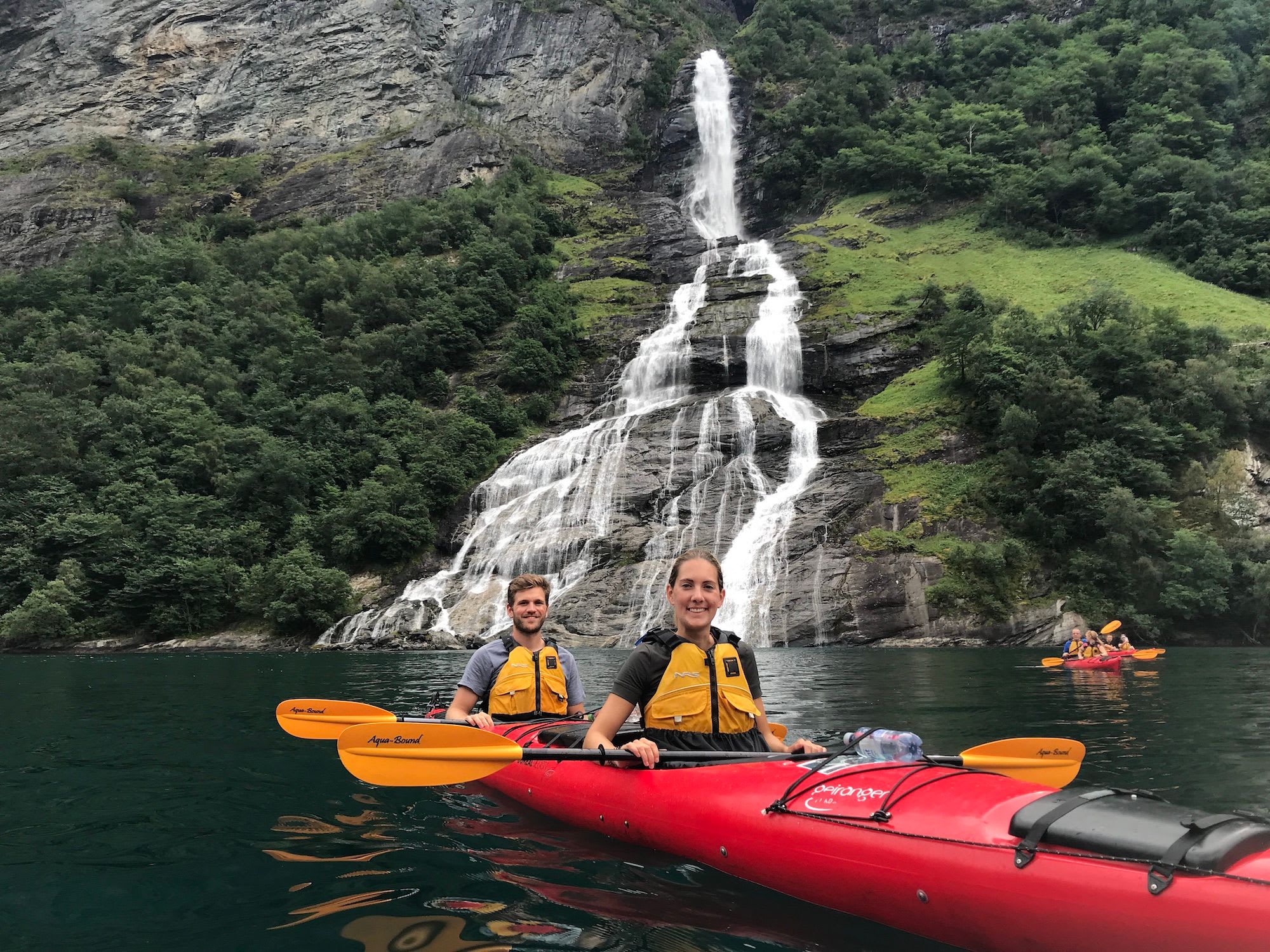 Mann og kvinne som padler i en rød kajakk med oransje padler, smilende til kameraet med et fossefall og grønn natur i bakgrunnen.