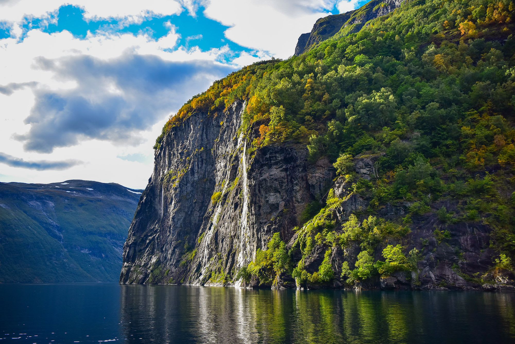 A cliff towering over a calm fjord with vibrant autumn colors reflected on the water.