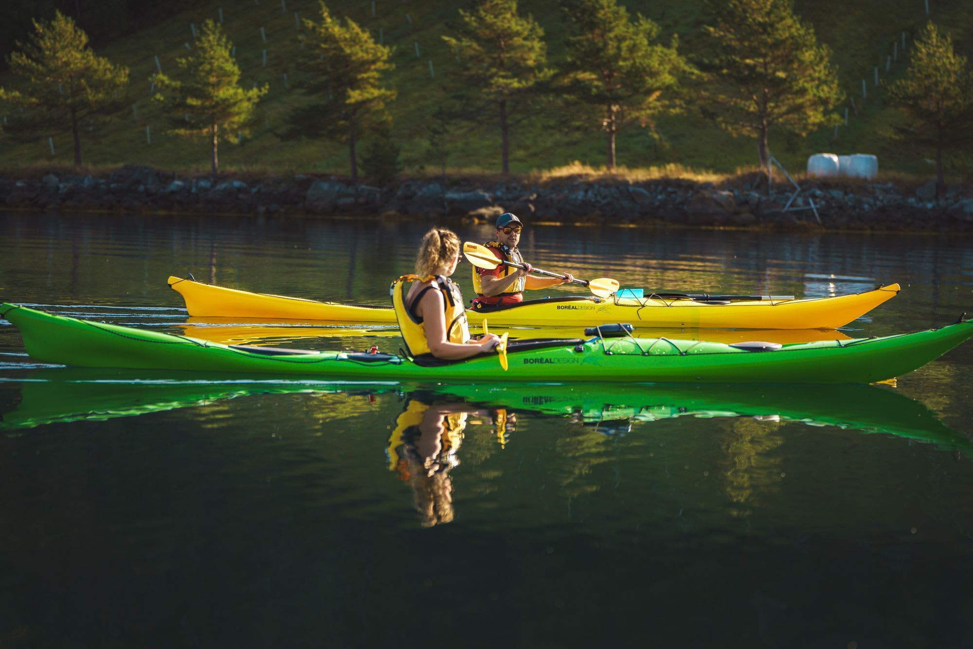 Kvinne og mann som padler side om side på rolig vann, med kajakker i grønn og gul, omringet av trær og en steinete strandlinje.