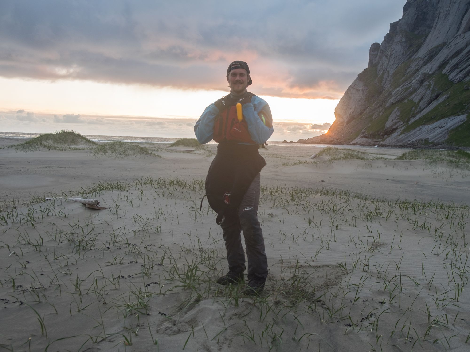 Smilende person på strand i kajakkutstyr, med solnedgang og hav.
