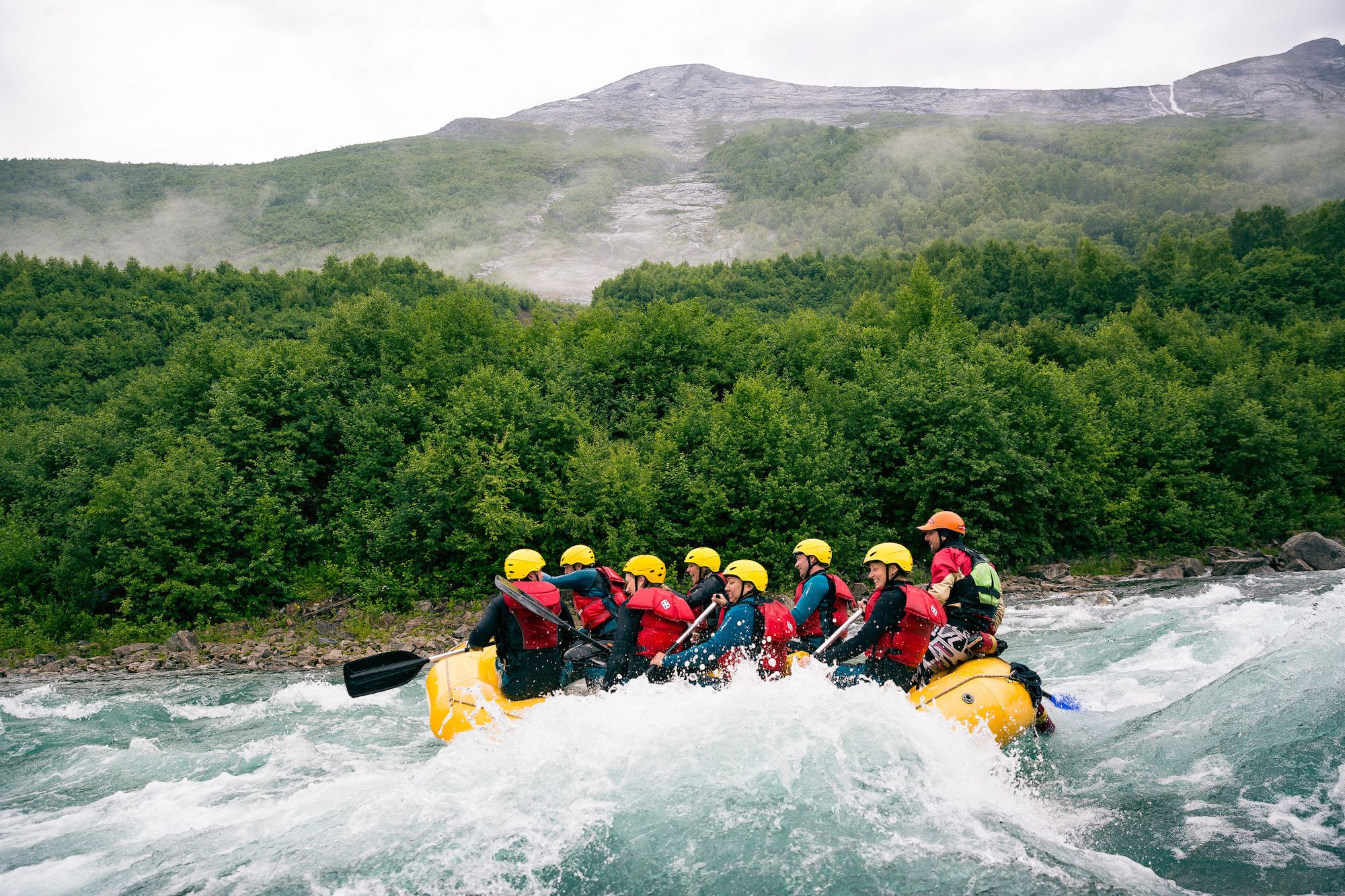 A group of people rafting through rough water in a yellow raft, wearing helmets and life jackets, surrounded by trees and mountains.