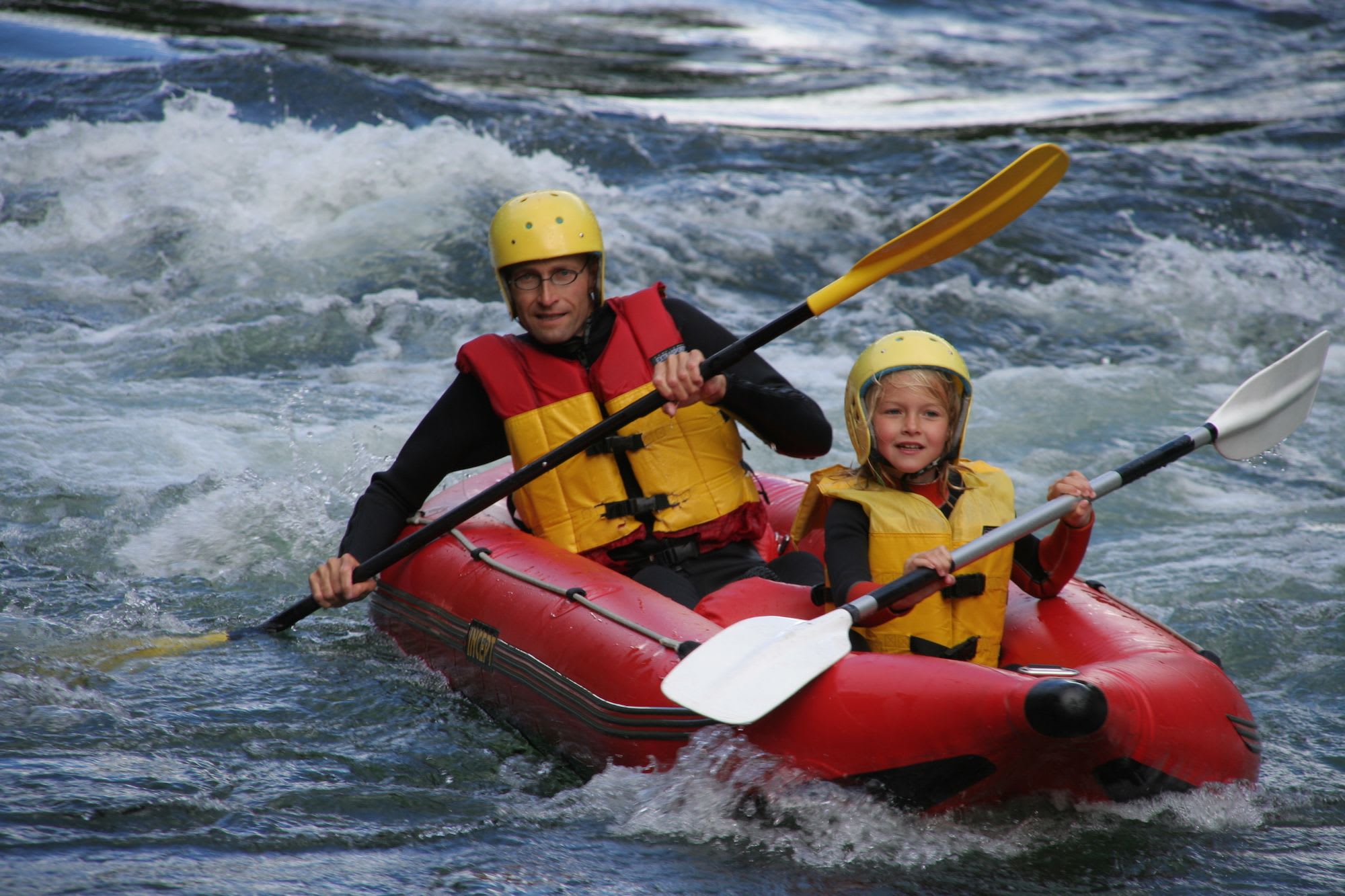 A father and daughter paddling together in a red inflatable boat through mild rapids.