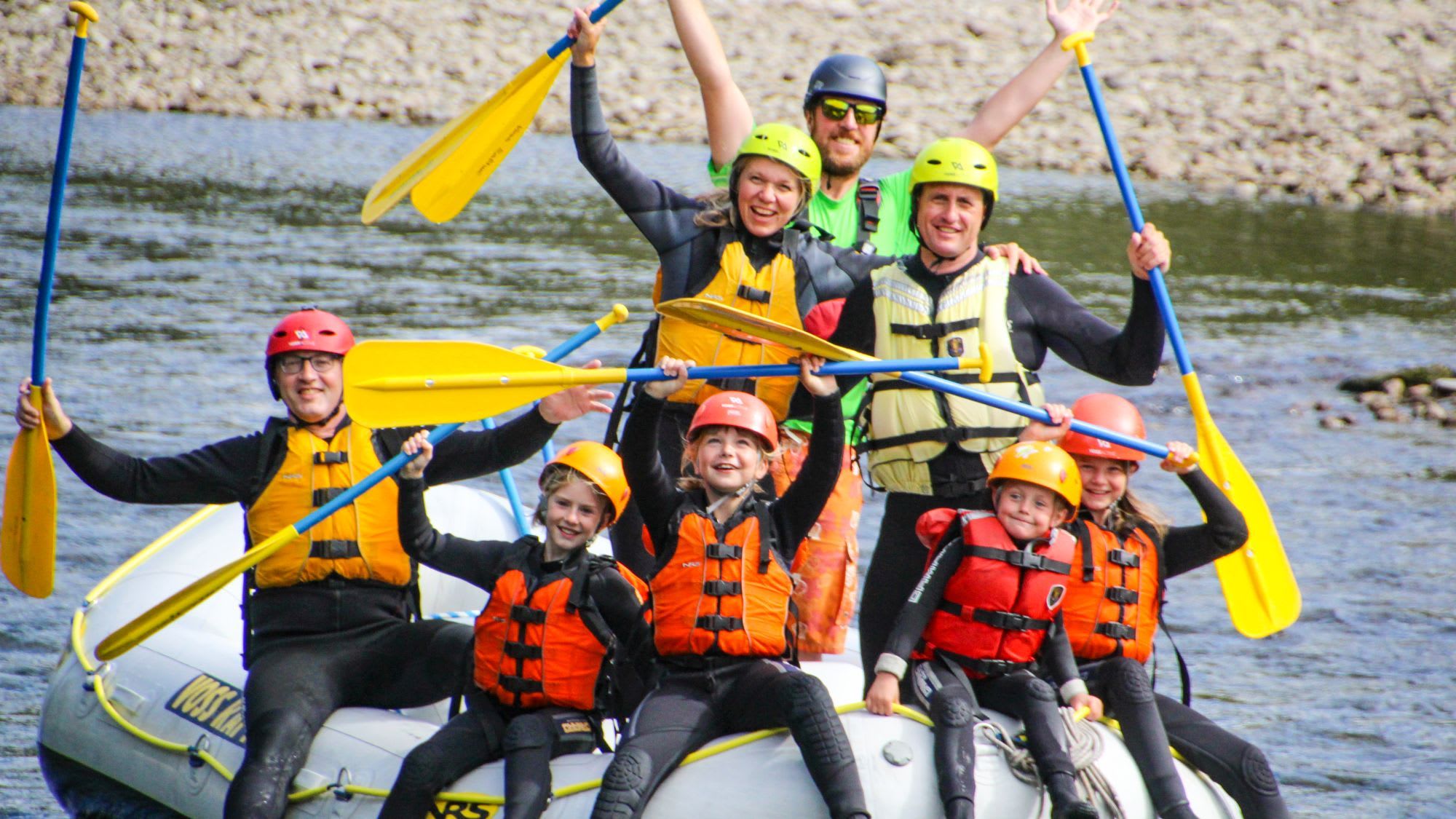 A group rafting down a calm river in a white raft, wearing bright helmets and life jackets, raising their arms in excitement.