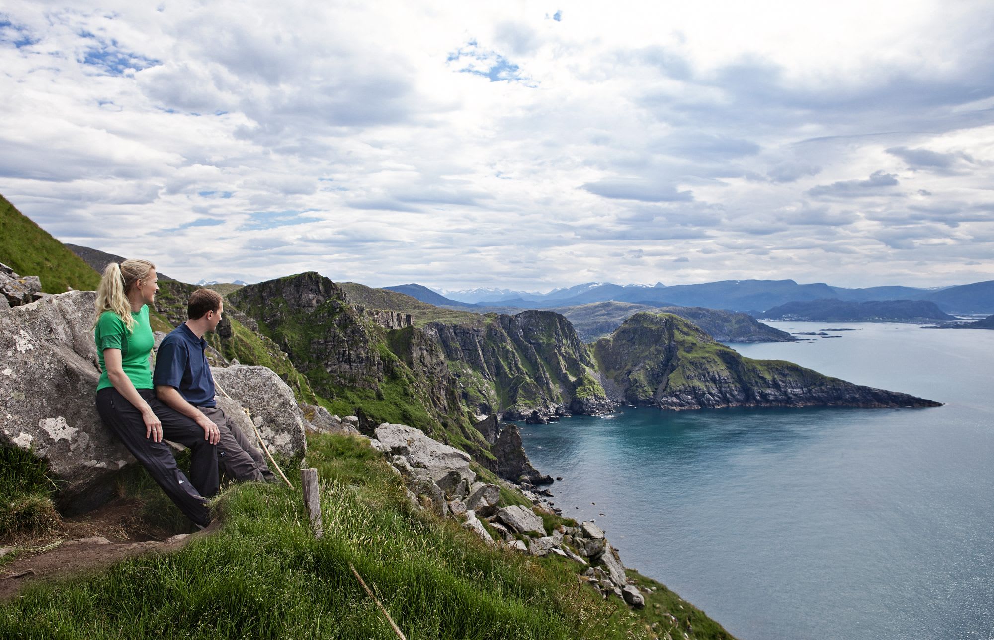 Et par sitter på store steiner og ser ut over et kystlandskap med klipper, bakker og hav under en delvis skyet himmel.