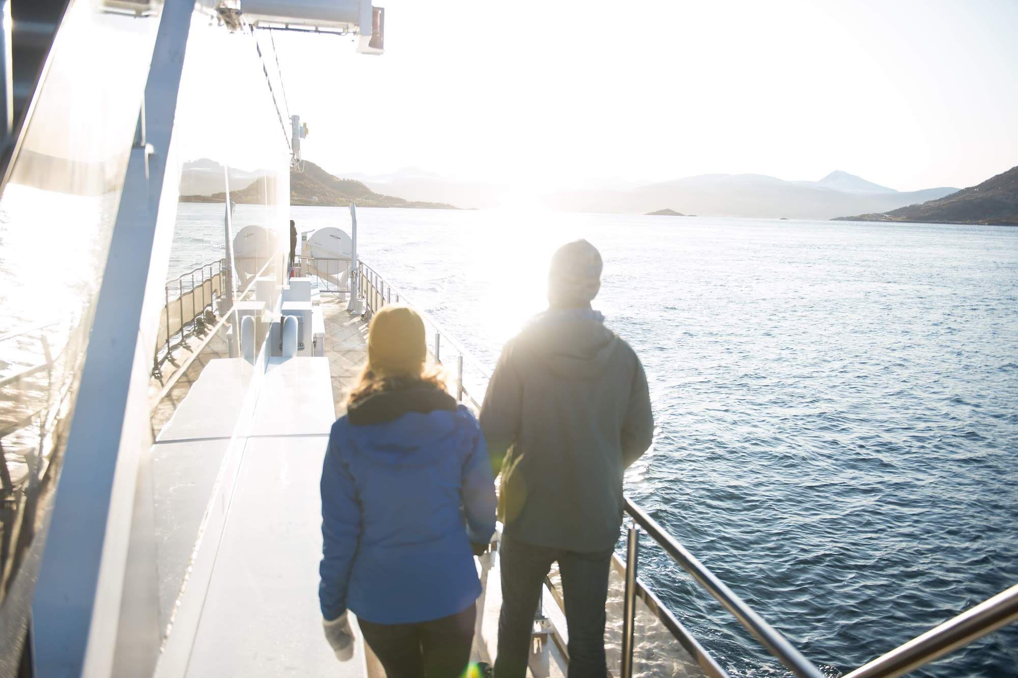 Two people walk along boat deck at sunset, gazing at calm waters.