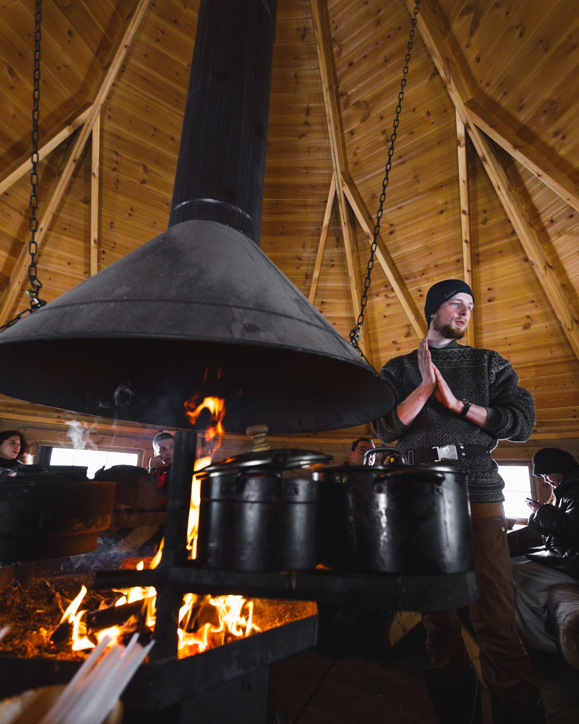 Warm cabin interior, man speaking beside firepit with pots, people seated around.