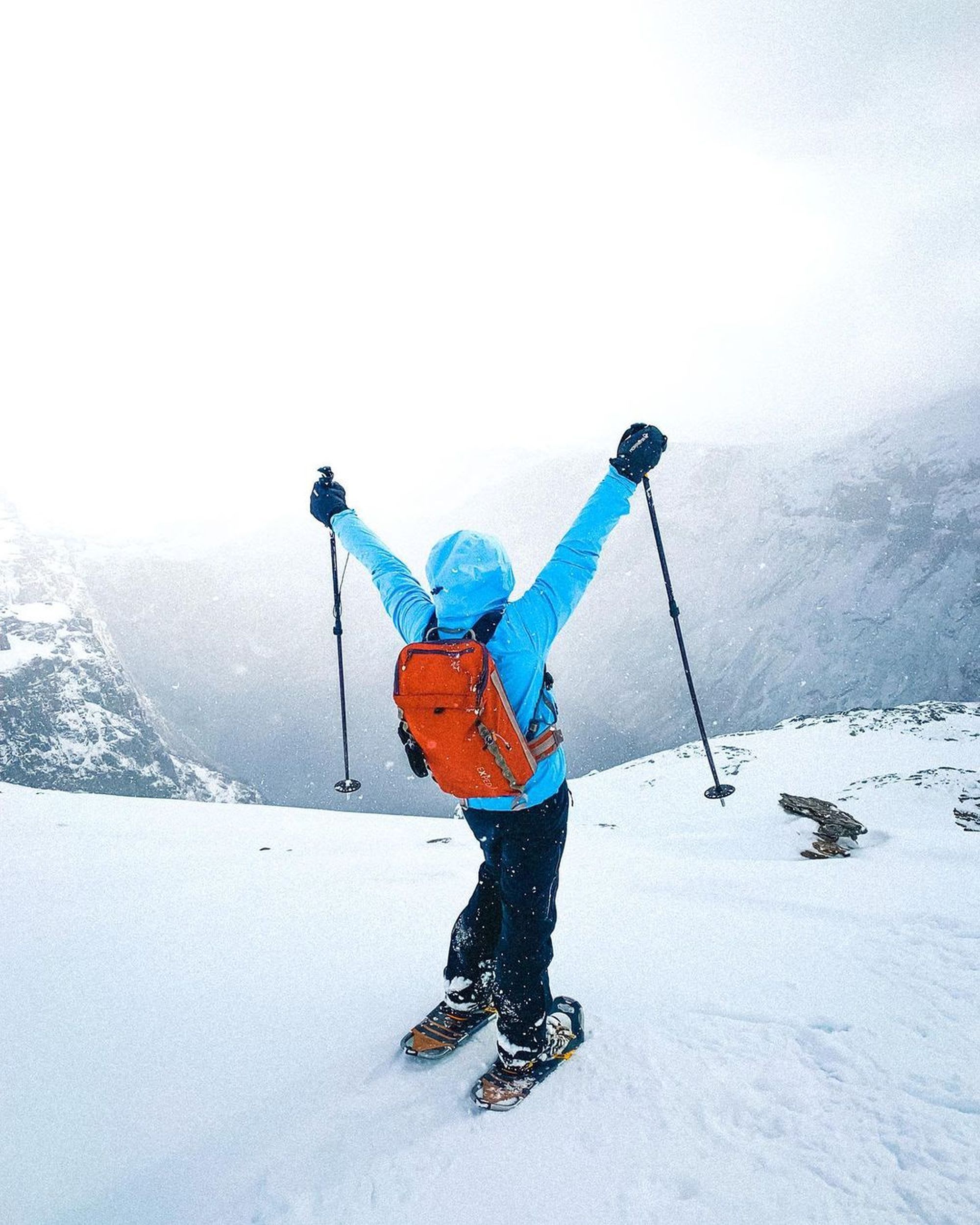 En person i blå jakke og oransje ryggsekk står triumferende i dyp snø med turstaver på en snødekt fjellrygg.