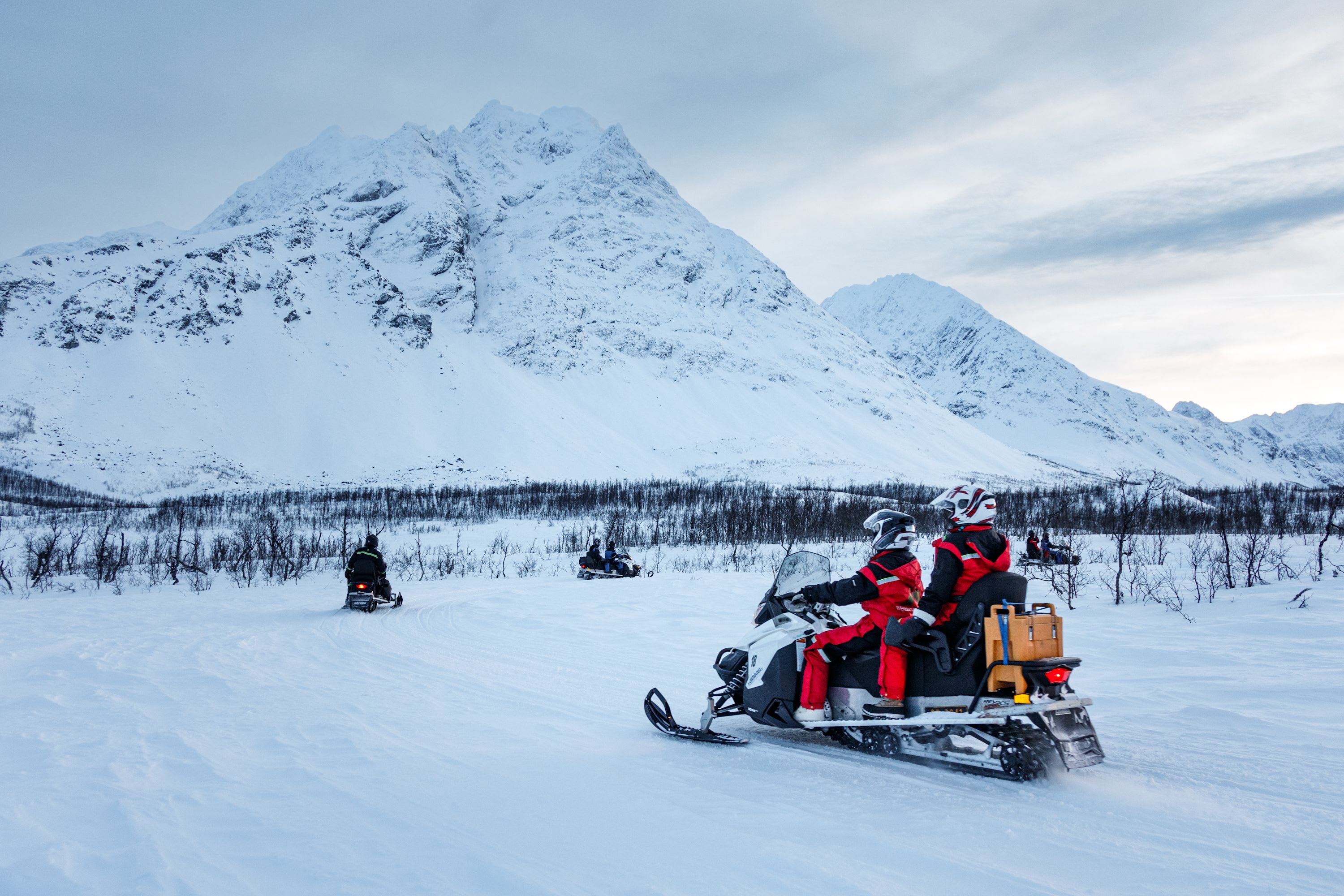 Flere snøscooterkjørere krysser åpent snølandskap med dramatiske fjelltopper i bakgrunnen.