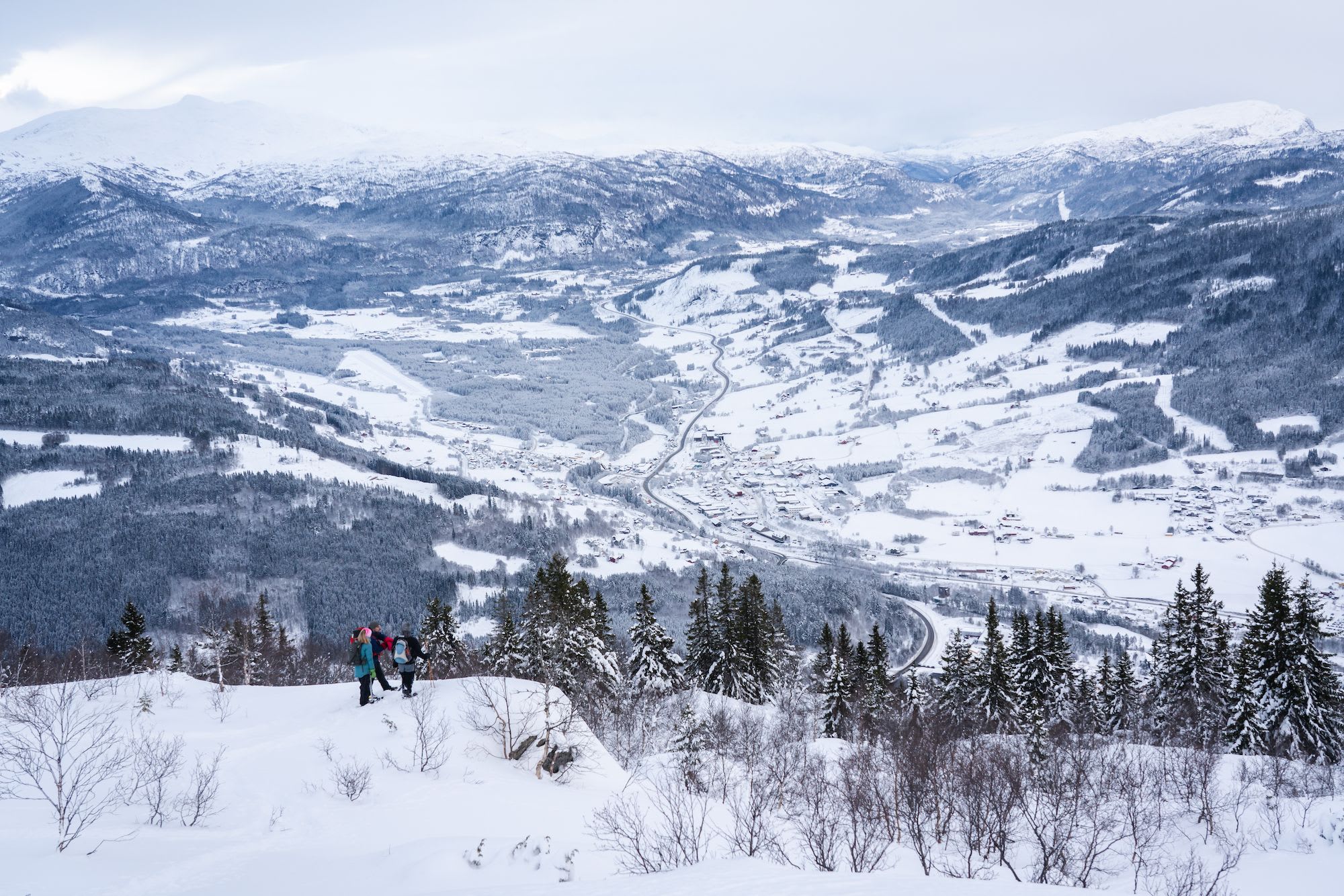 Three hikers walk through deep snow on mountain above snowy valley.
