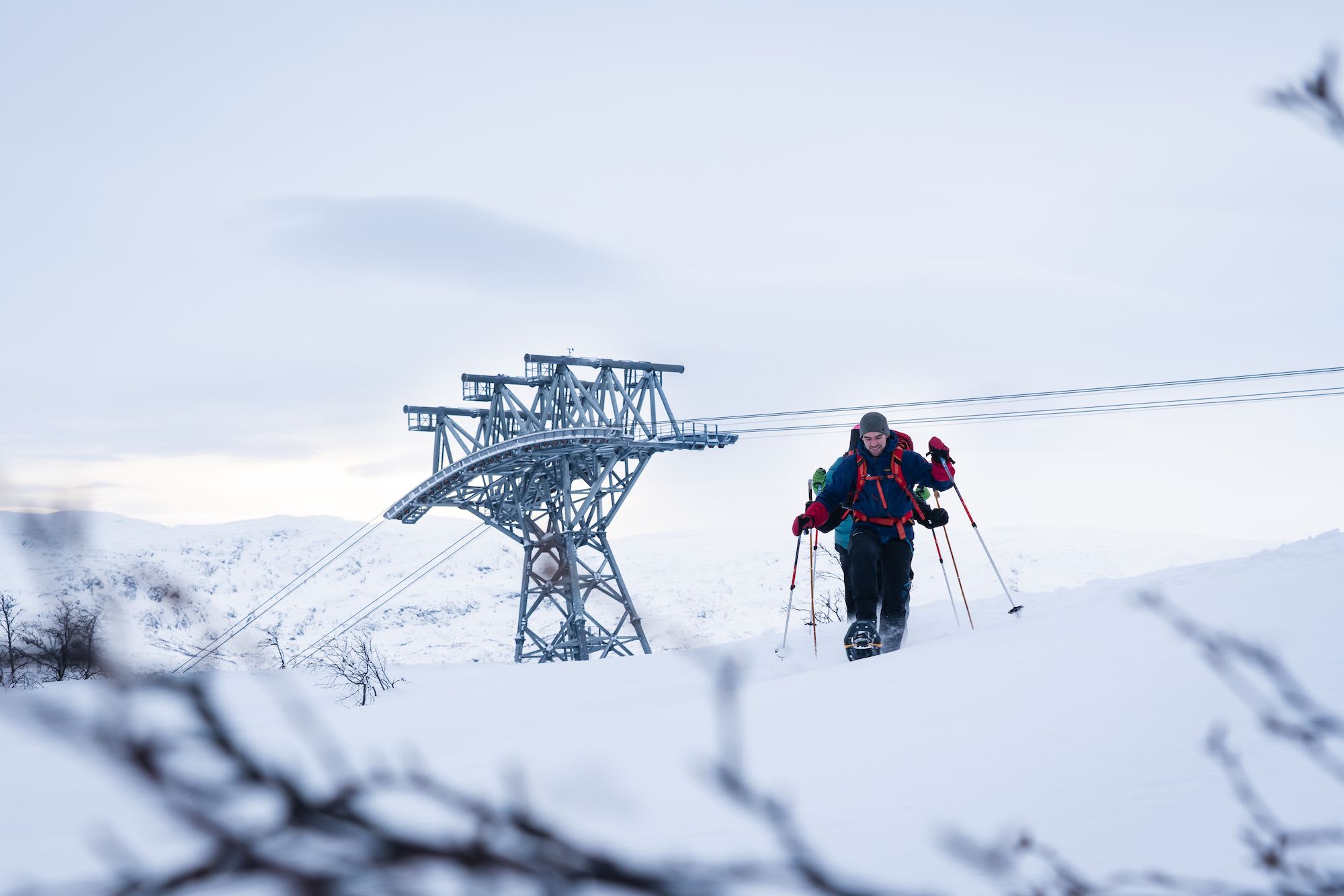 Two snowshoers climb past gondola tower in snowy landscape.