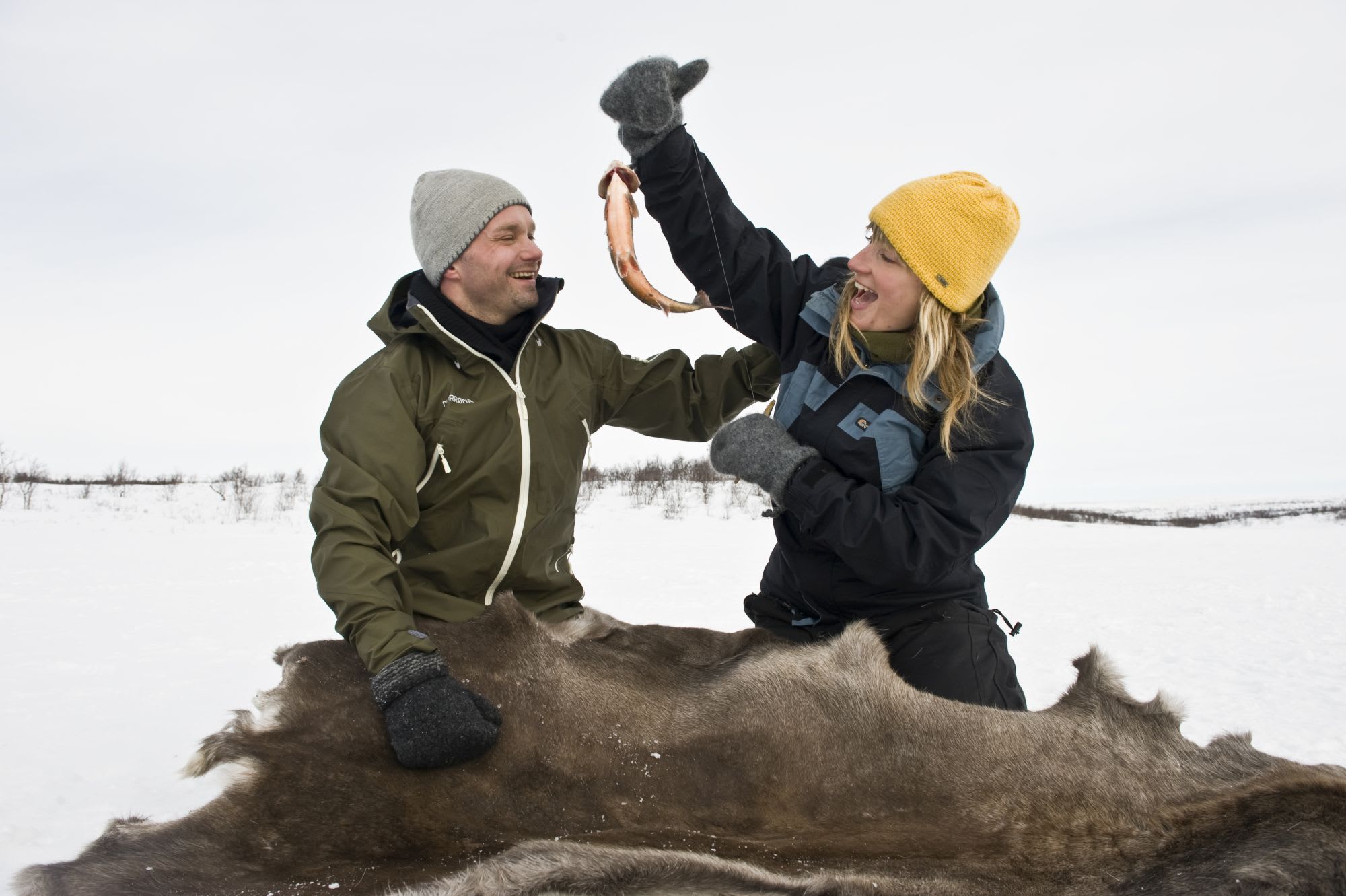 To Isfiskere sitter på et reinsdyrskinn i snøen og ler glad, mens en av dem holder opp en nyfanget fisk.