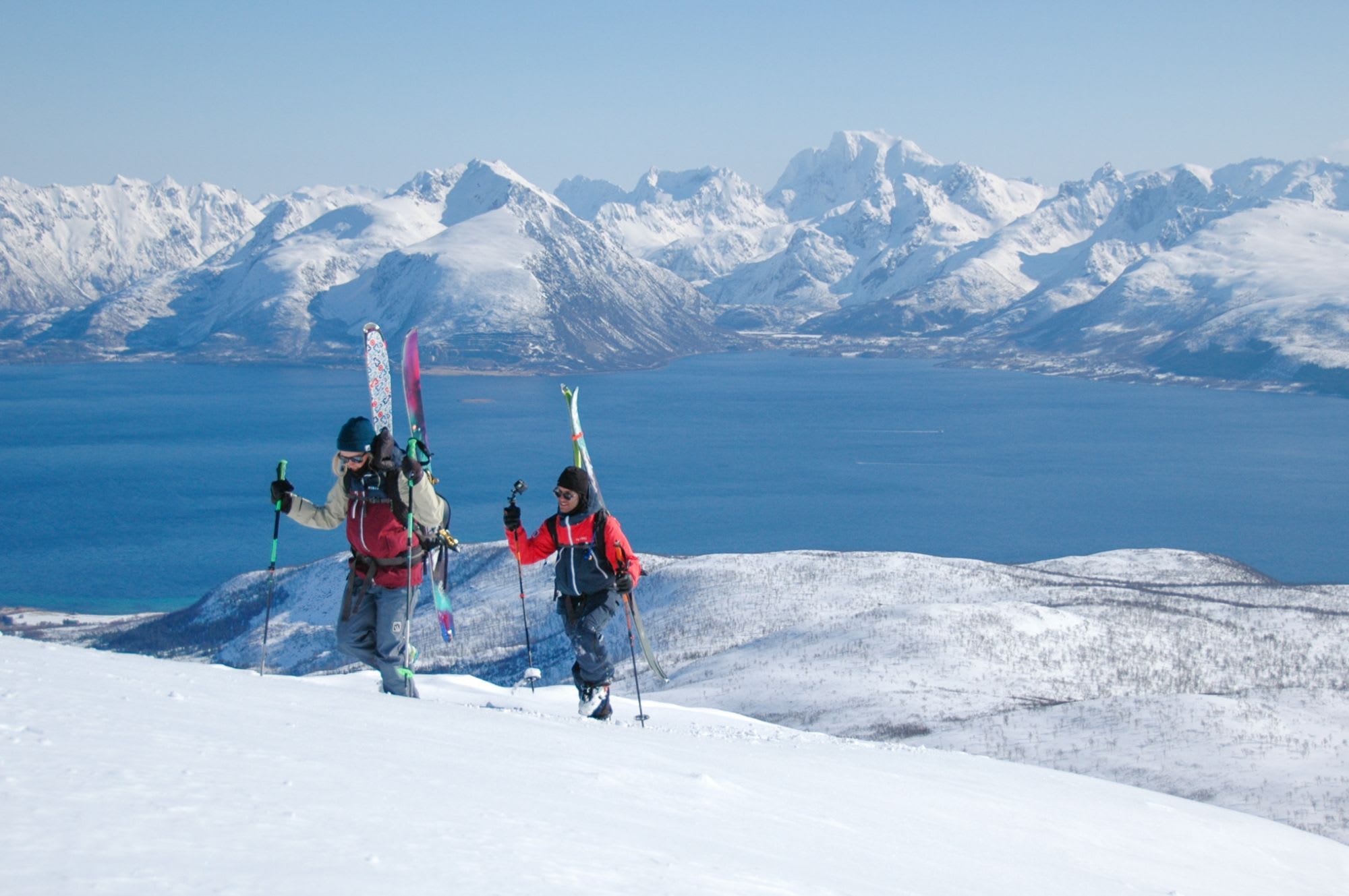 Two skiers hike uphill through snow with skis on their backpacks, surrounded by snowy mountains and a deep blue fjord.