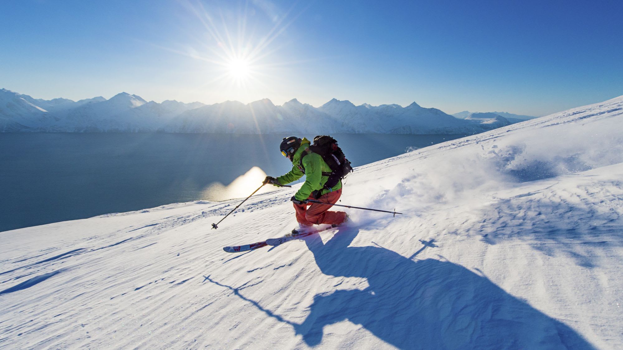 A skier in bright gear glides down a sunlit snowy slope, casting a long shadow, with a calm fjord and rugged mountains in the background.