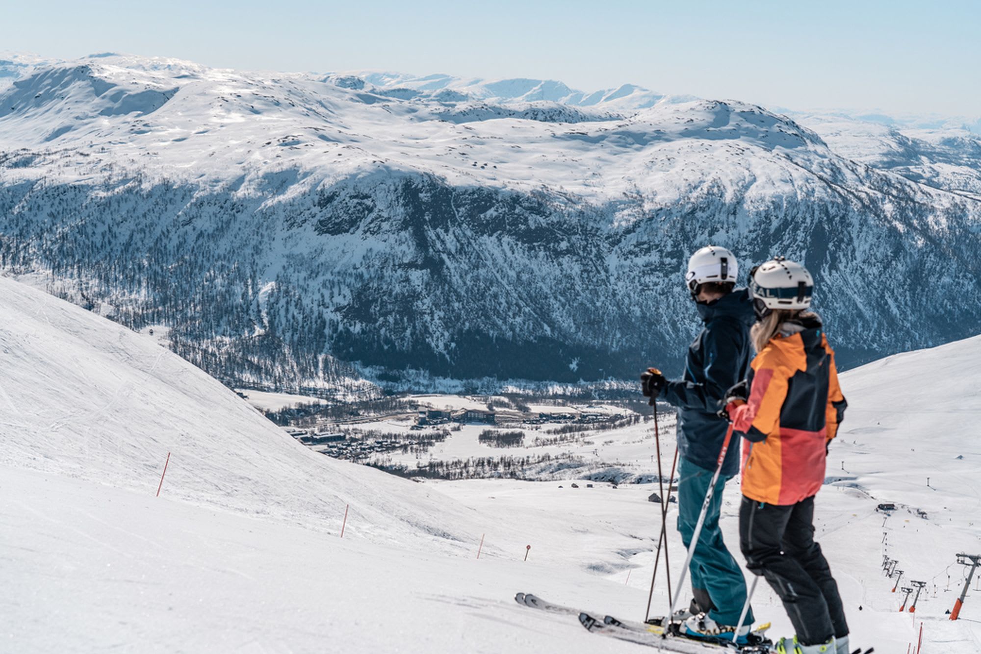 Zwei Skifahrer stehen auf einer schneebedeckten Piste und blicken auf die Berglandschaft mit Skiliften in der Ferne.