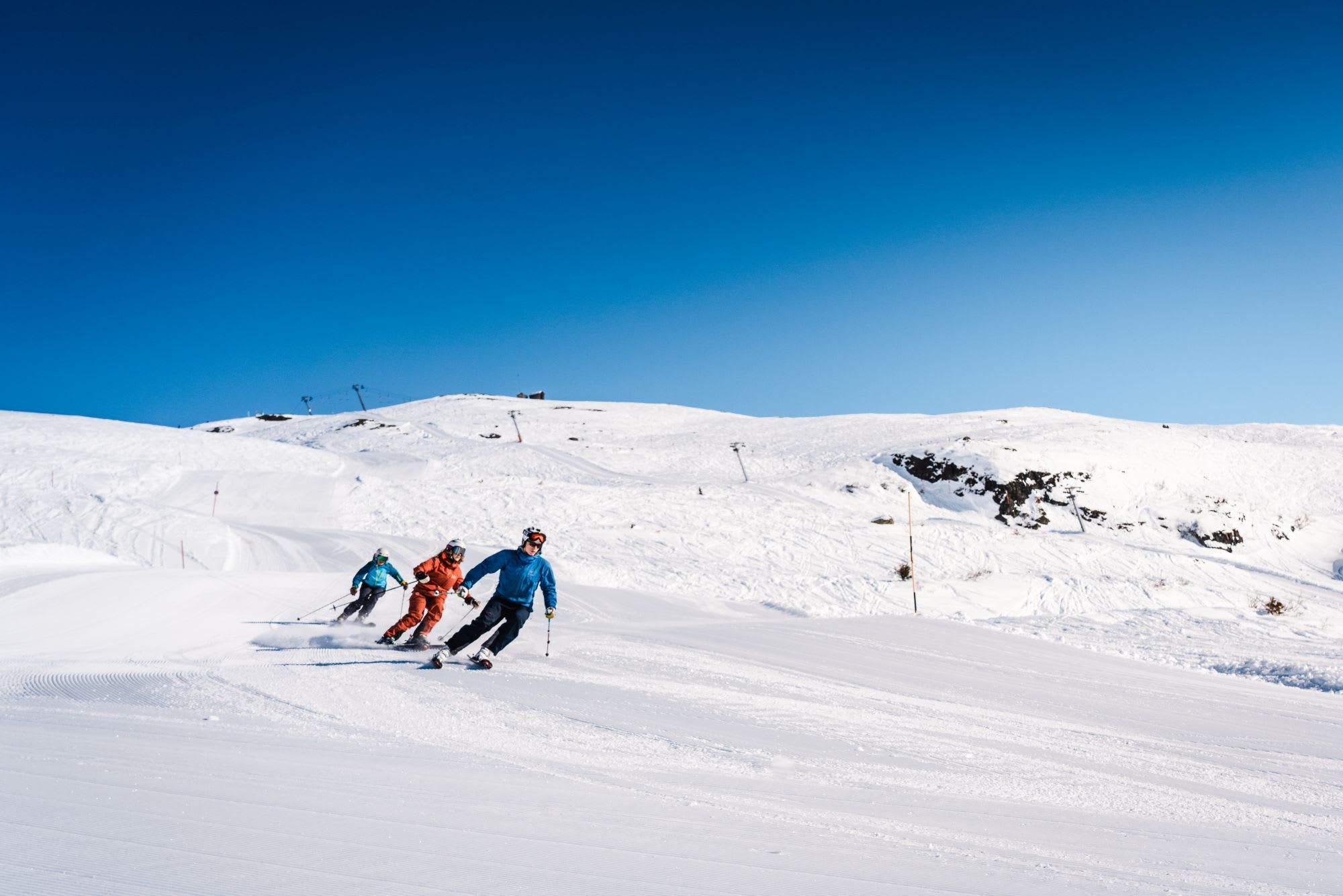 A group of three skiers carving down a snow-covered slope under a clear blue sky, with ski lifts and mountains in the background.