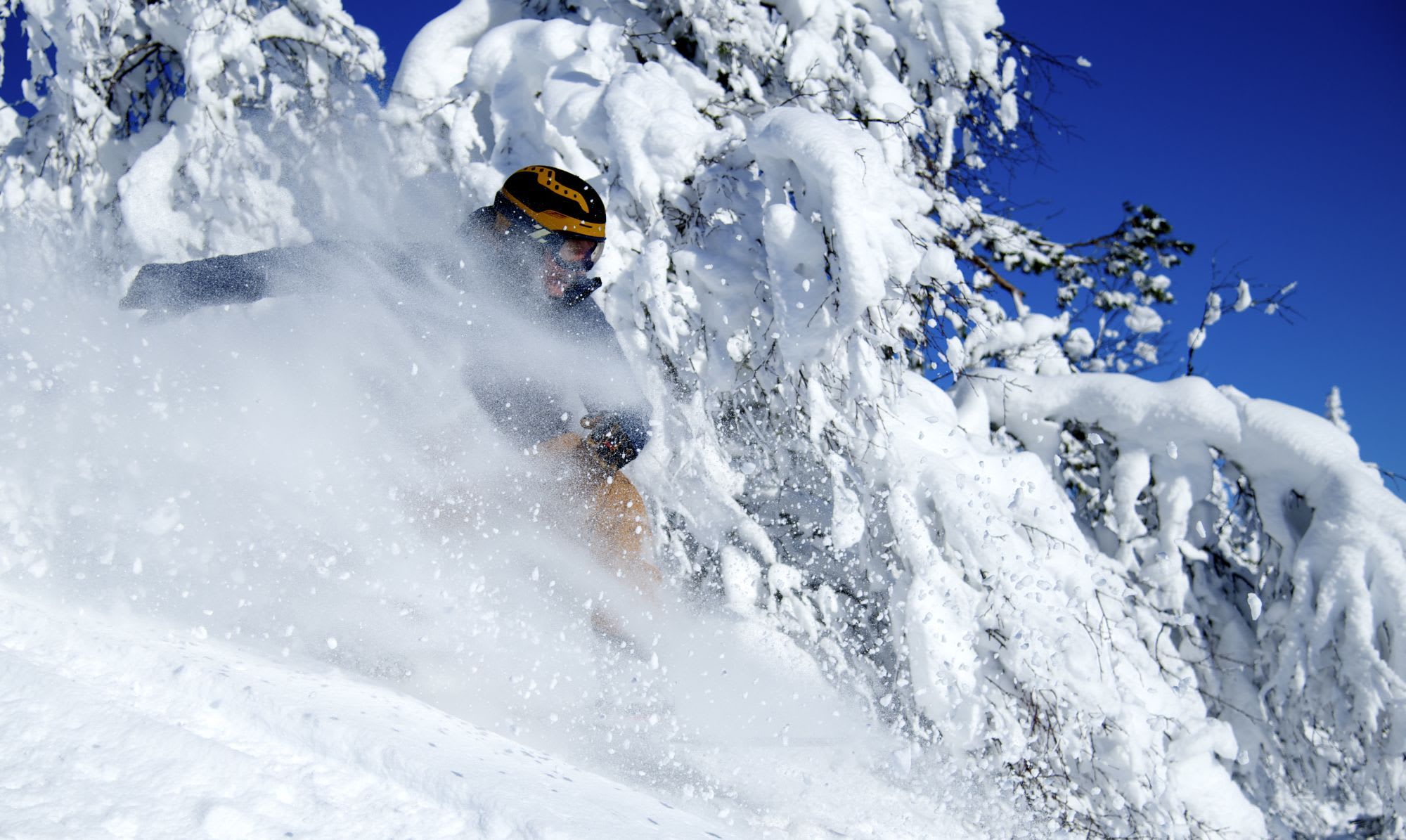 A skier in a yellow helmet and goggles carves through deep powder snow, kicking up a flurry of snowflakes.
