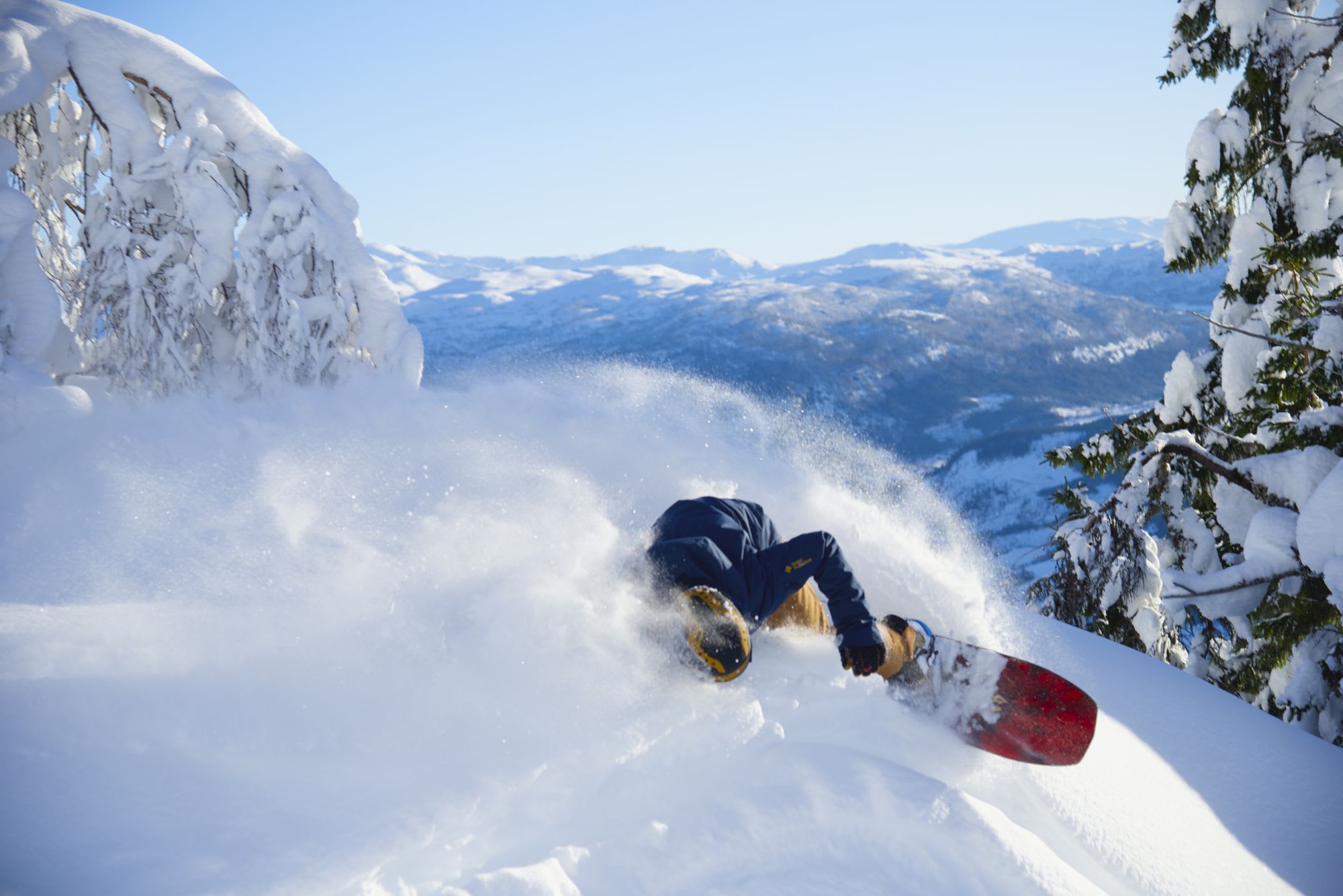 A snowboarder in a blue jacket and yellow helmet carving through deep snow, sending up a cloud of snow spray.
