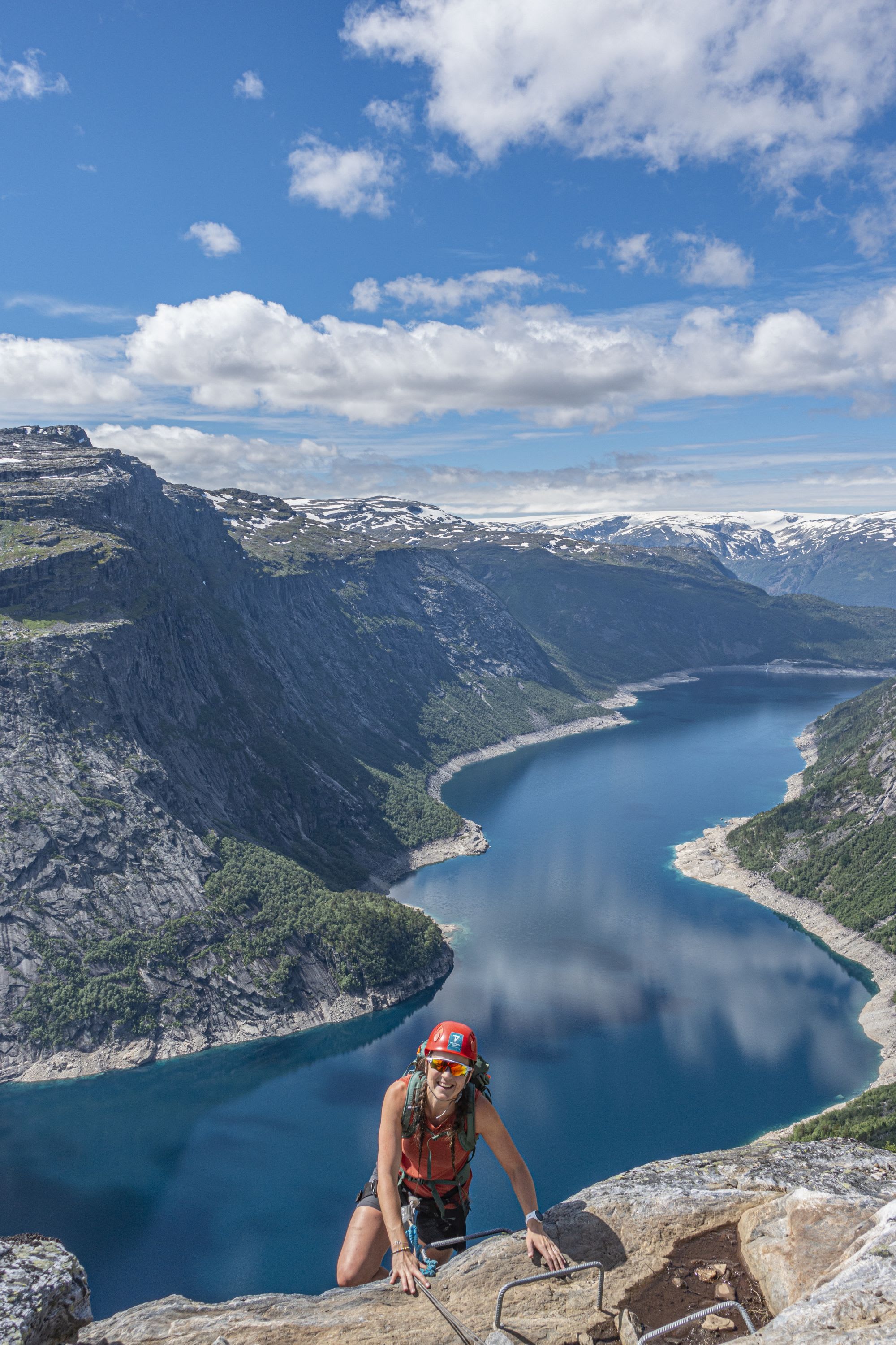 Climber in helmet and sunglasses smiles while scaling cliff over fjord.