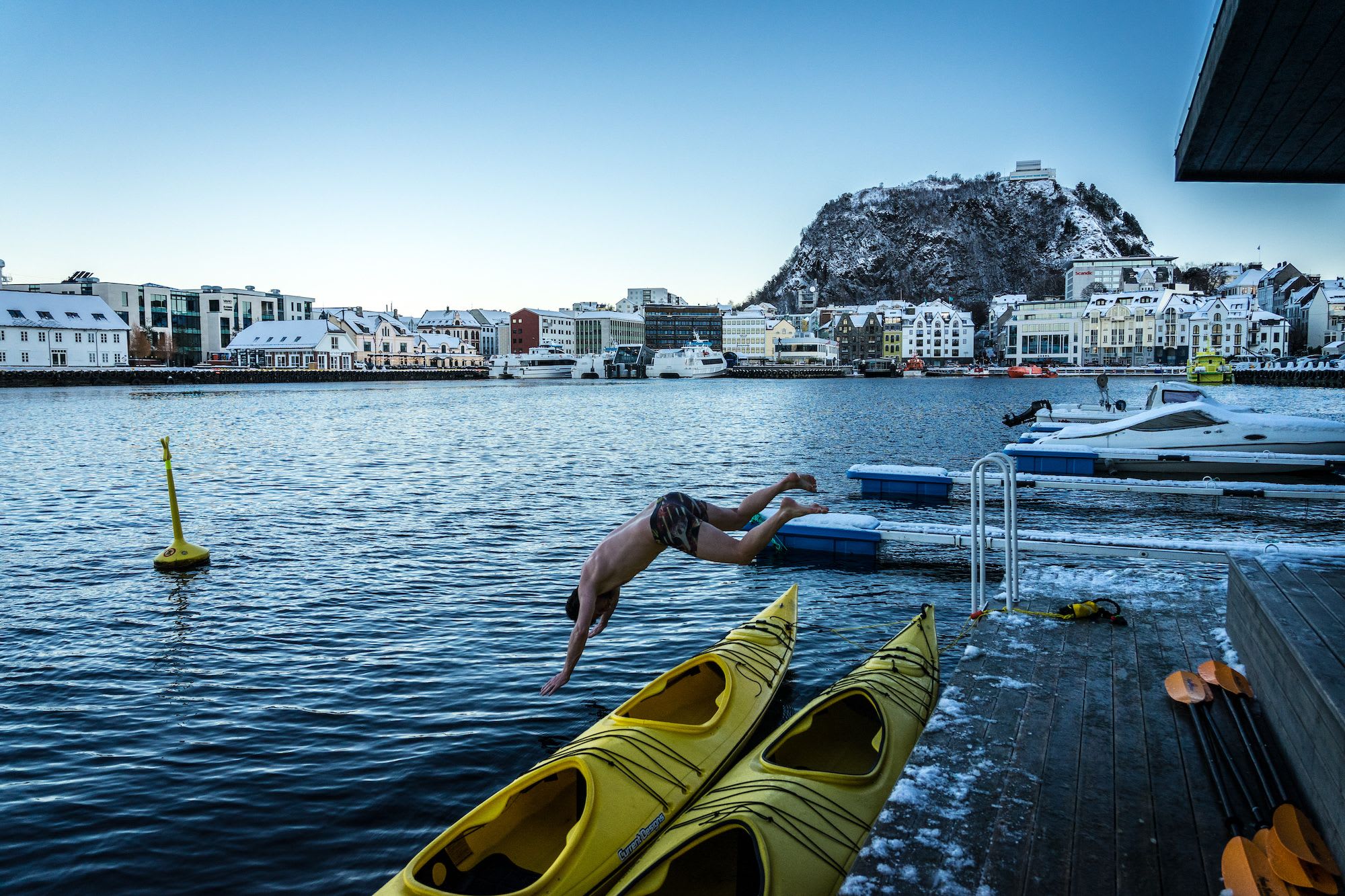 Eine Person springt in kaltes Wasser von einem Dock, mit schneebedeckten Gebäuden und einem Berg im Hintergrund.
