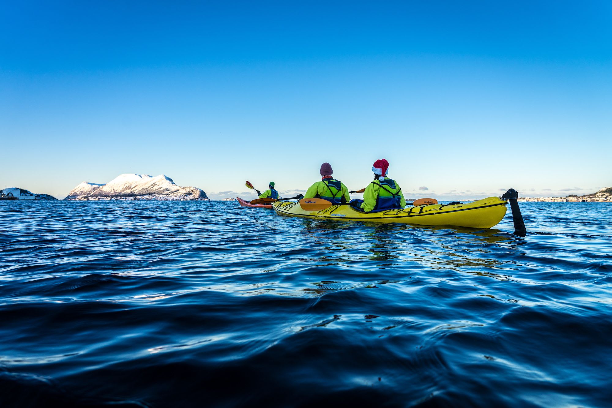 Drei Personen paddeln, wobei eine einen Weihnachtsmann-Hut trägt, auf dem Wasser mit schneebedeckten Bergen und klarem blauen Himmel im Hintergrund.