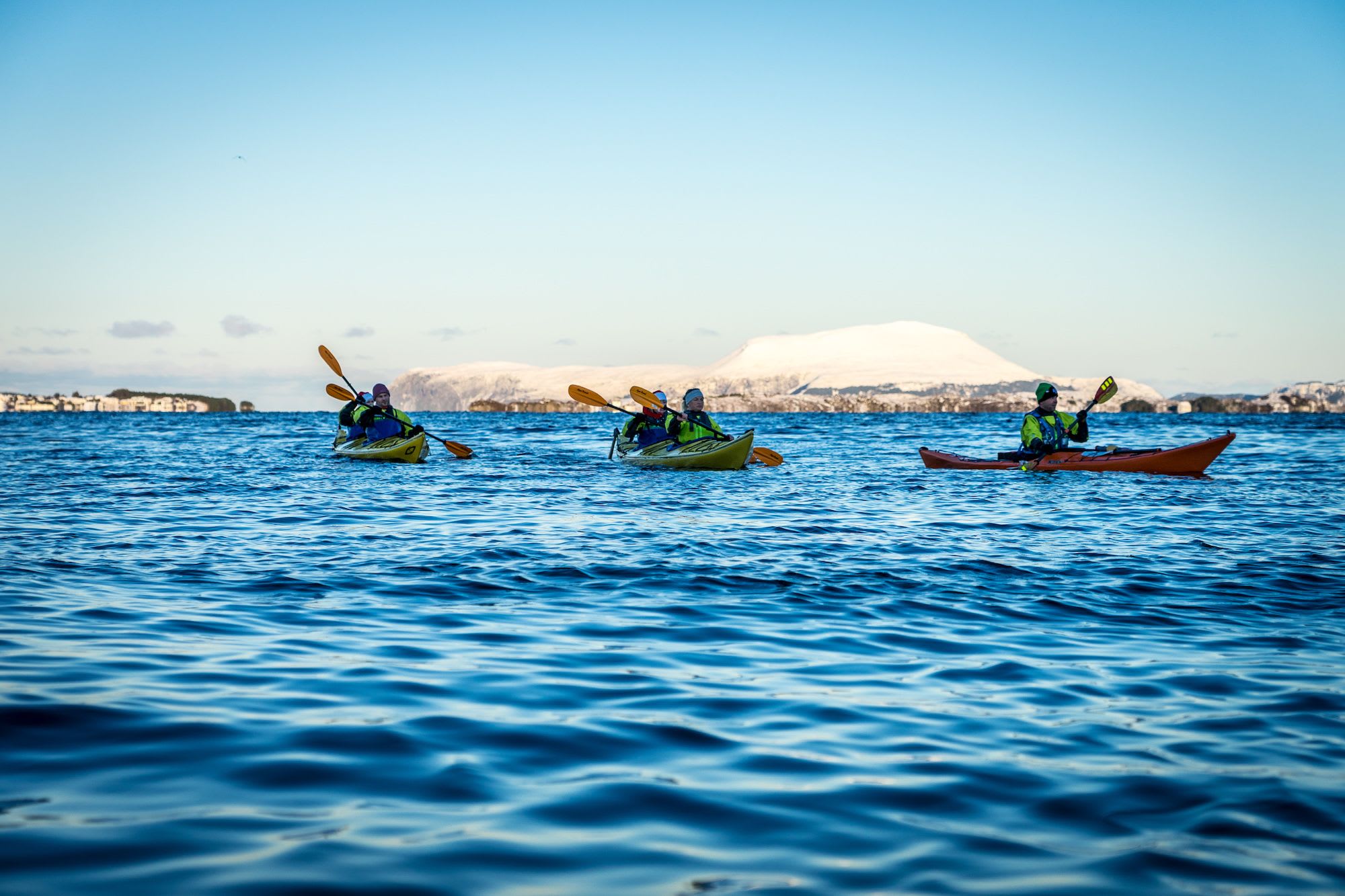 Fünf Personen paddeln auf ruhigem blauen Wasser, umgeben von schneebedeckten Bergen und Gebäuden entlang des Ufers.