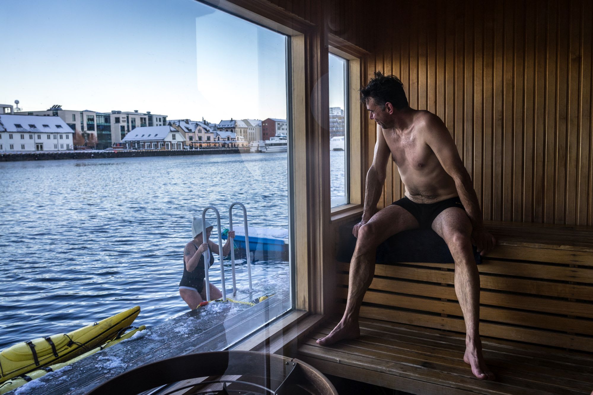 Ein Mann in Badebekleidung sitzt in einer Holzbadehaus und schaut einer Frau zu, die eine schneebedeckte Leiter aus dem Hafenwasser hinaufklettert.