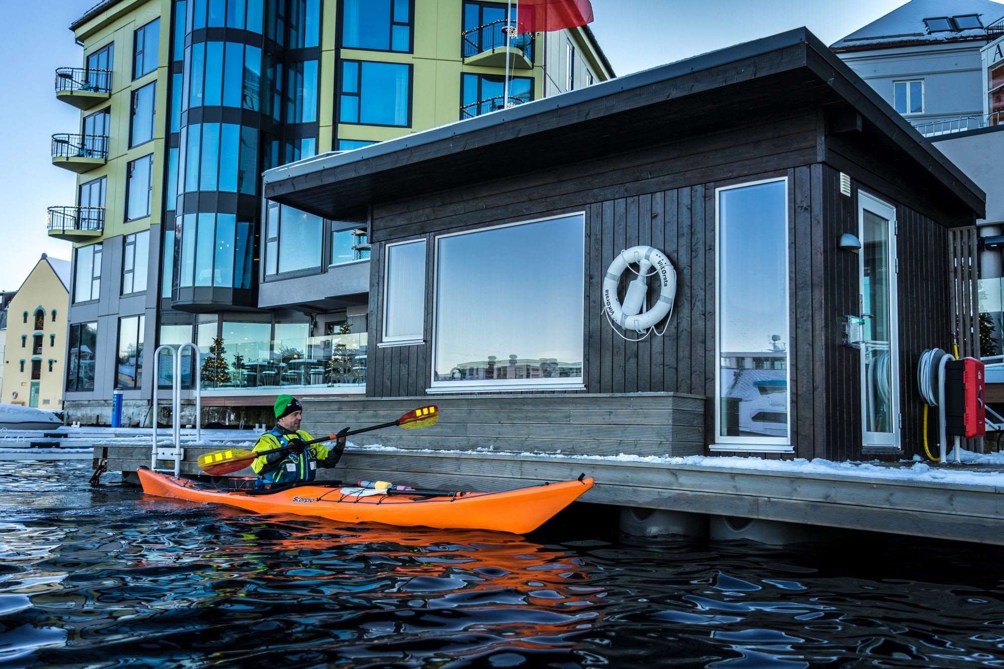 Eine Person mit grüner Mütze paddelt in einem orangefarbenen Kajak an einer modernen Sauna auf einem schneebedeckten Dock entlang, mit einem Hotel und Weihnachtsdekorationen im Hintergrund.