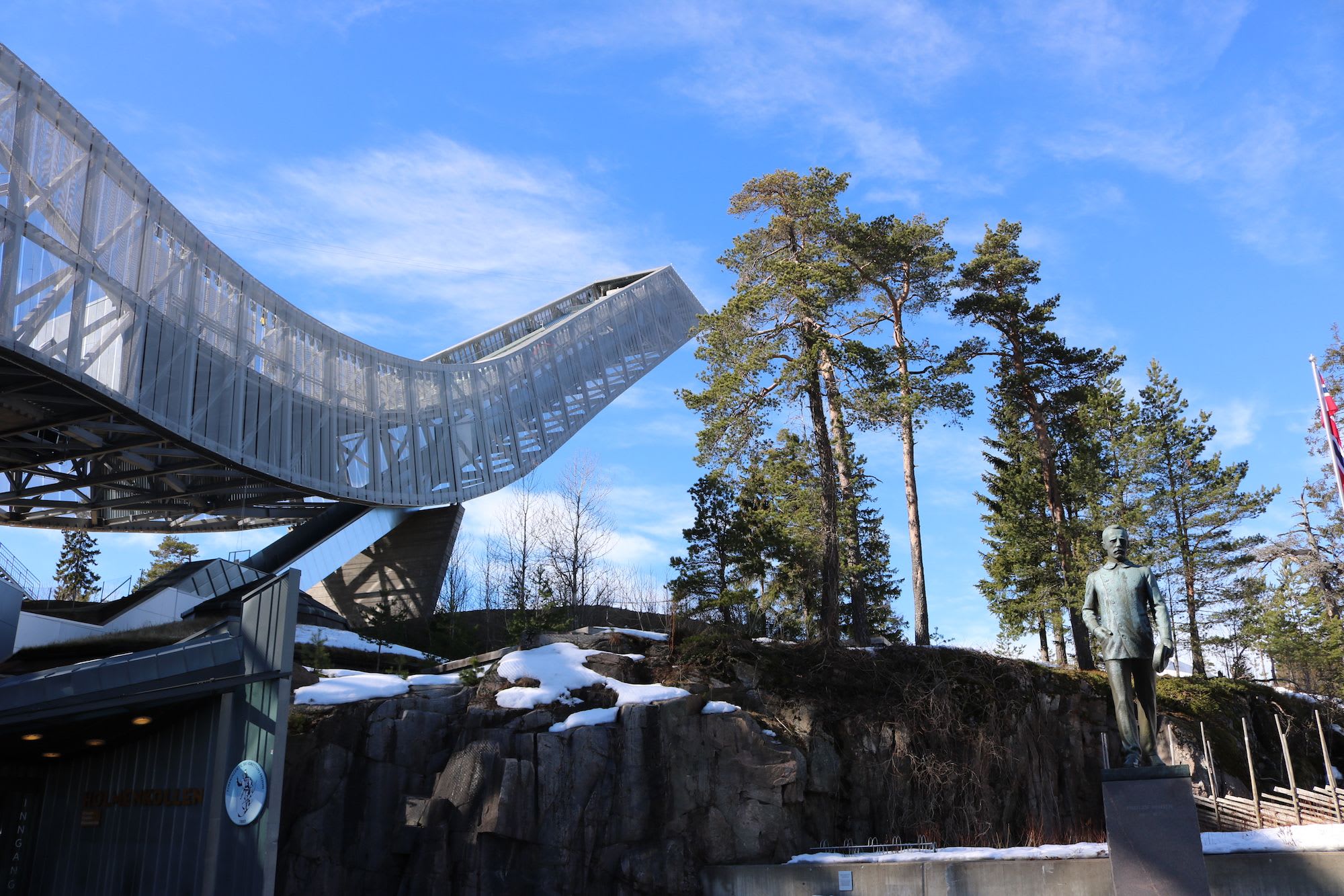 Nærbilde av Holmenkollen med statue foran og snødekte trær under blå himmel.