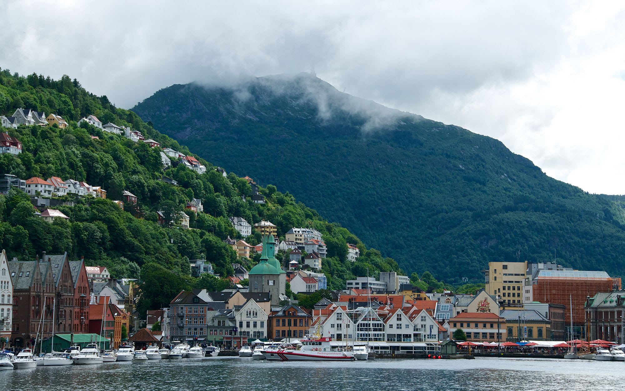 Scenic view of Bergen harbor with historic buildings, boats, and misty green hills rising behind.