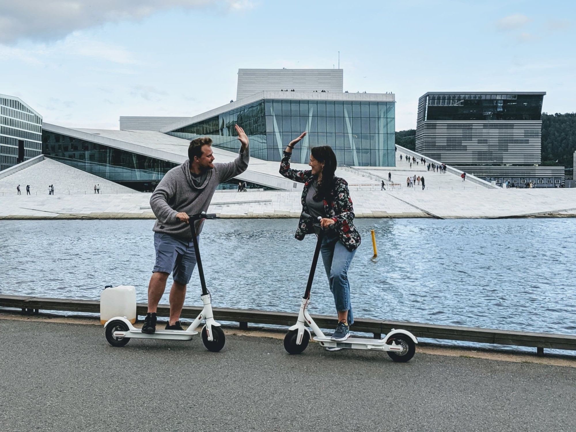 Man and woman on electric scooters high-five in front of Oslo Opera House by the water.