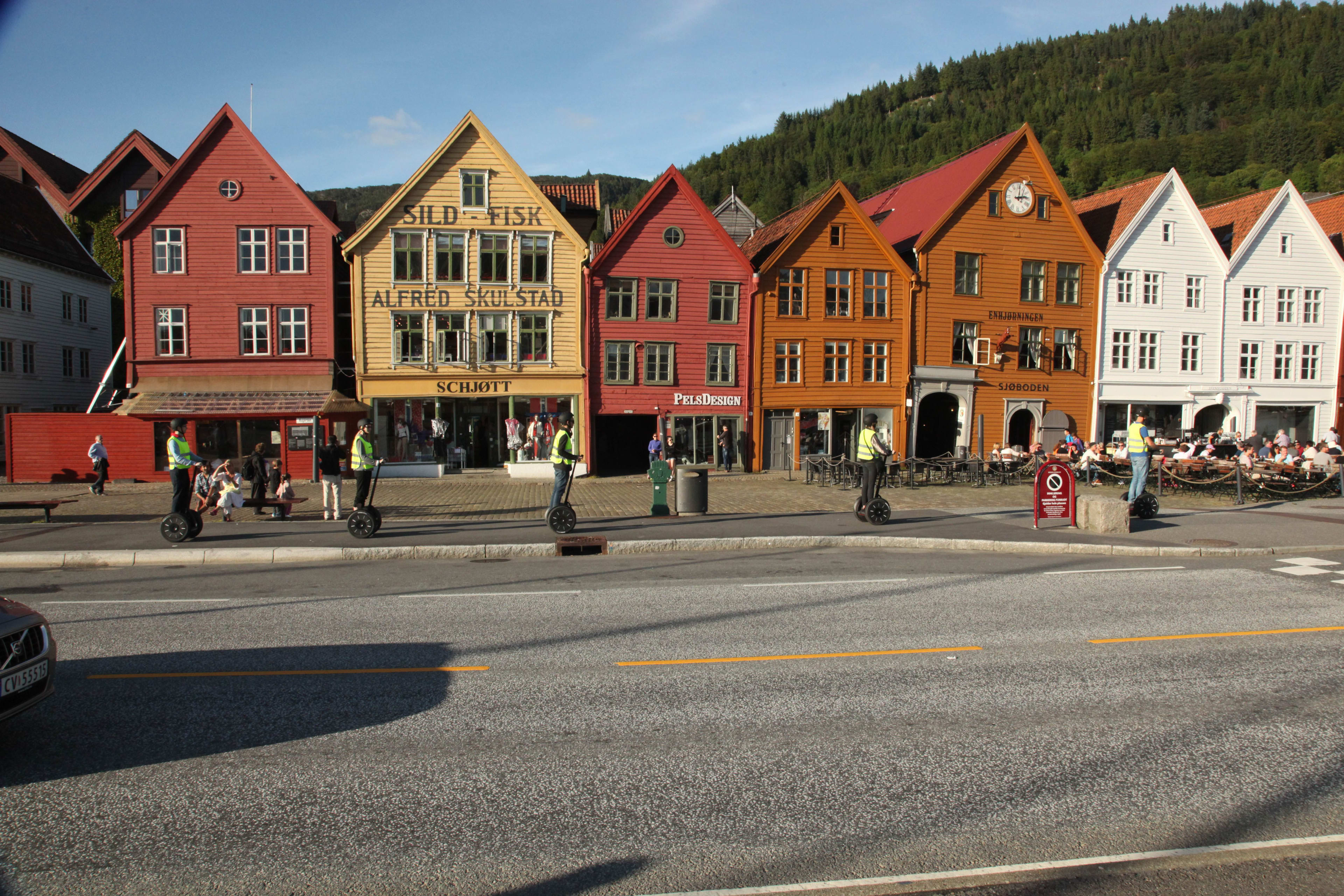 Segway riders cruise along Bryggen waterfront in Bergen under a clear sky, passing historic wooden buildings.