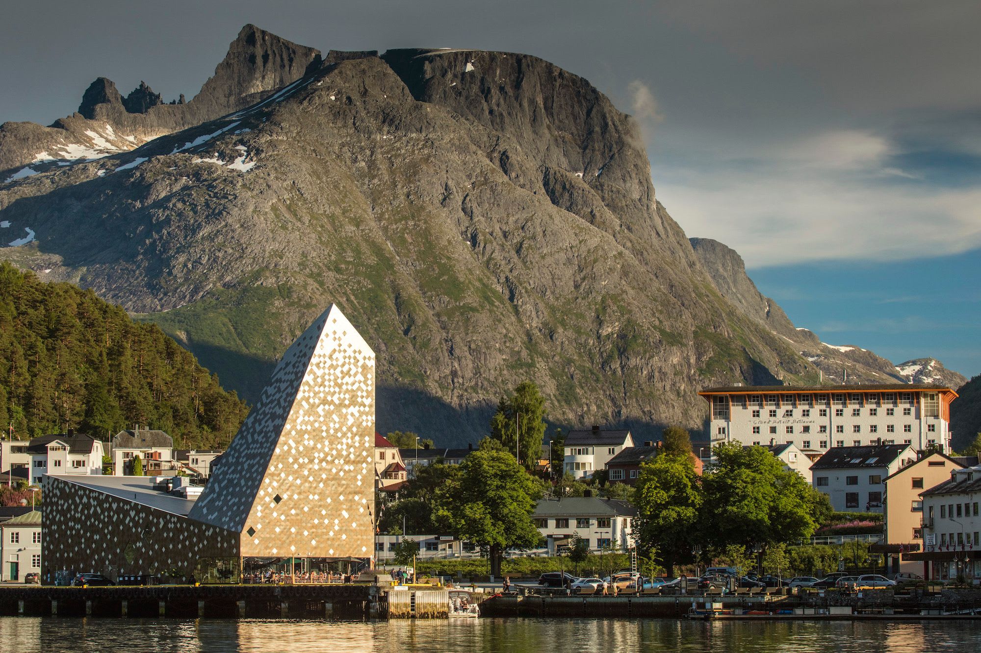 En moderne bygning med en geometrisk fasade, plassert nær en brygge med bratte, snødekte fjell i bakgrunnen.
